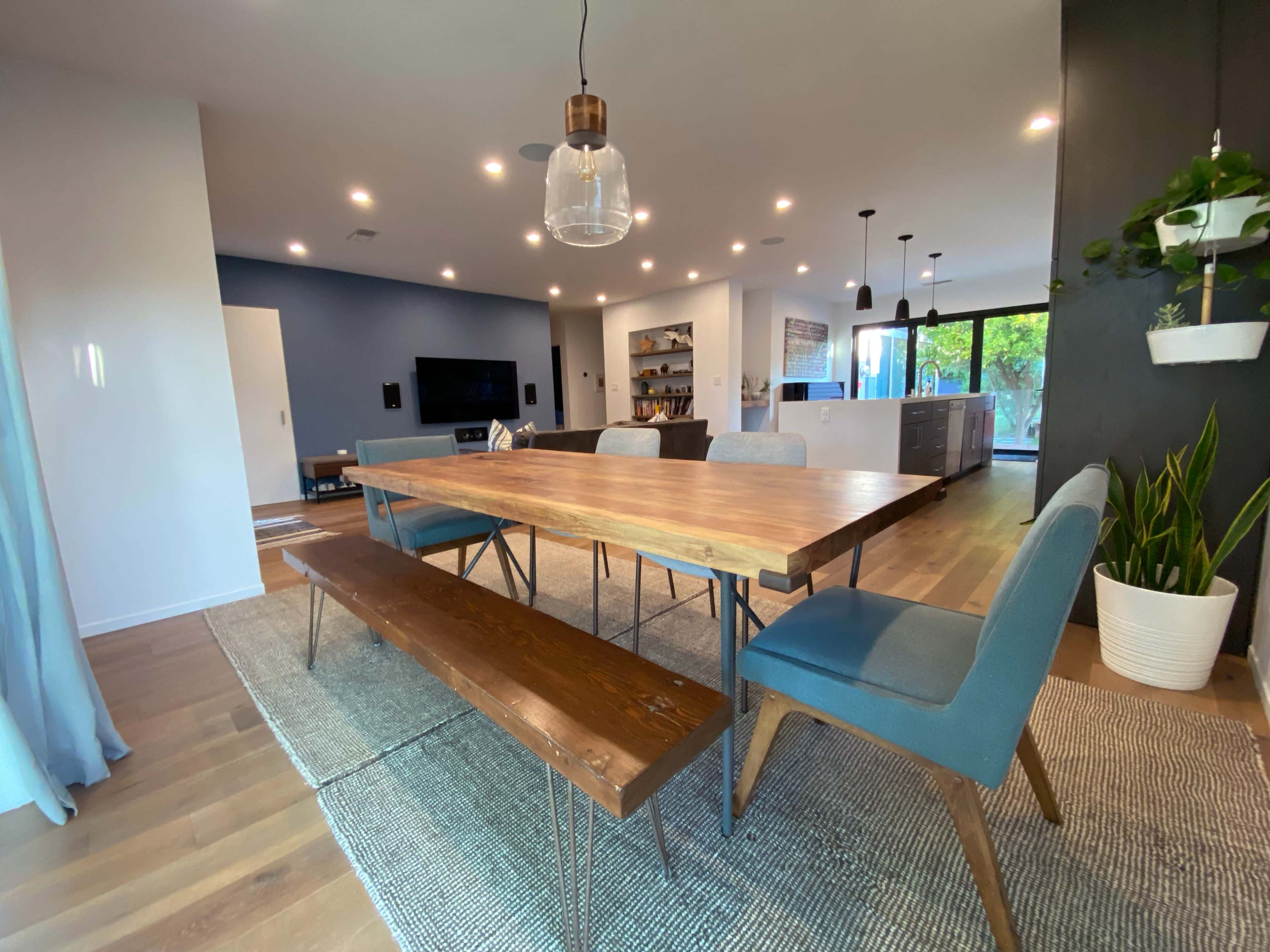 A modern dining room with a wooden table, blue chairs, and a bench, surrounded by large windows and contemporary decor.