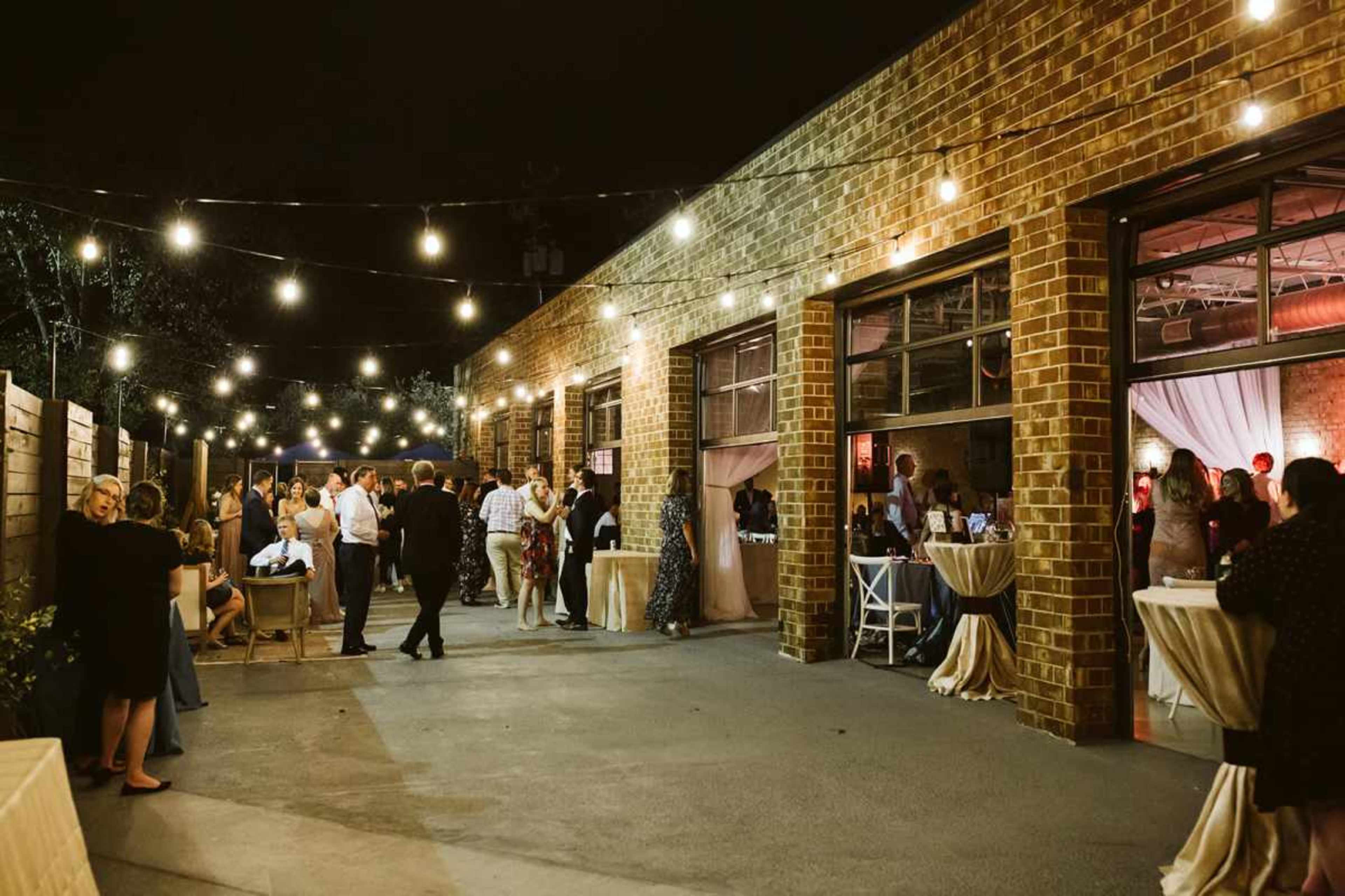 The image shows an outdoor evening event with string lights illuminating a gathering of people mingling around tables near a brick venue.