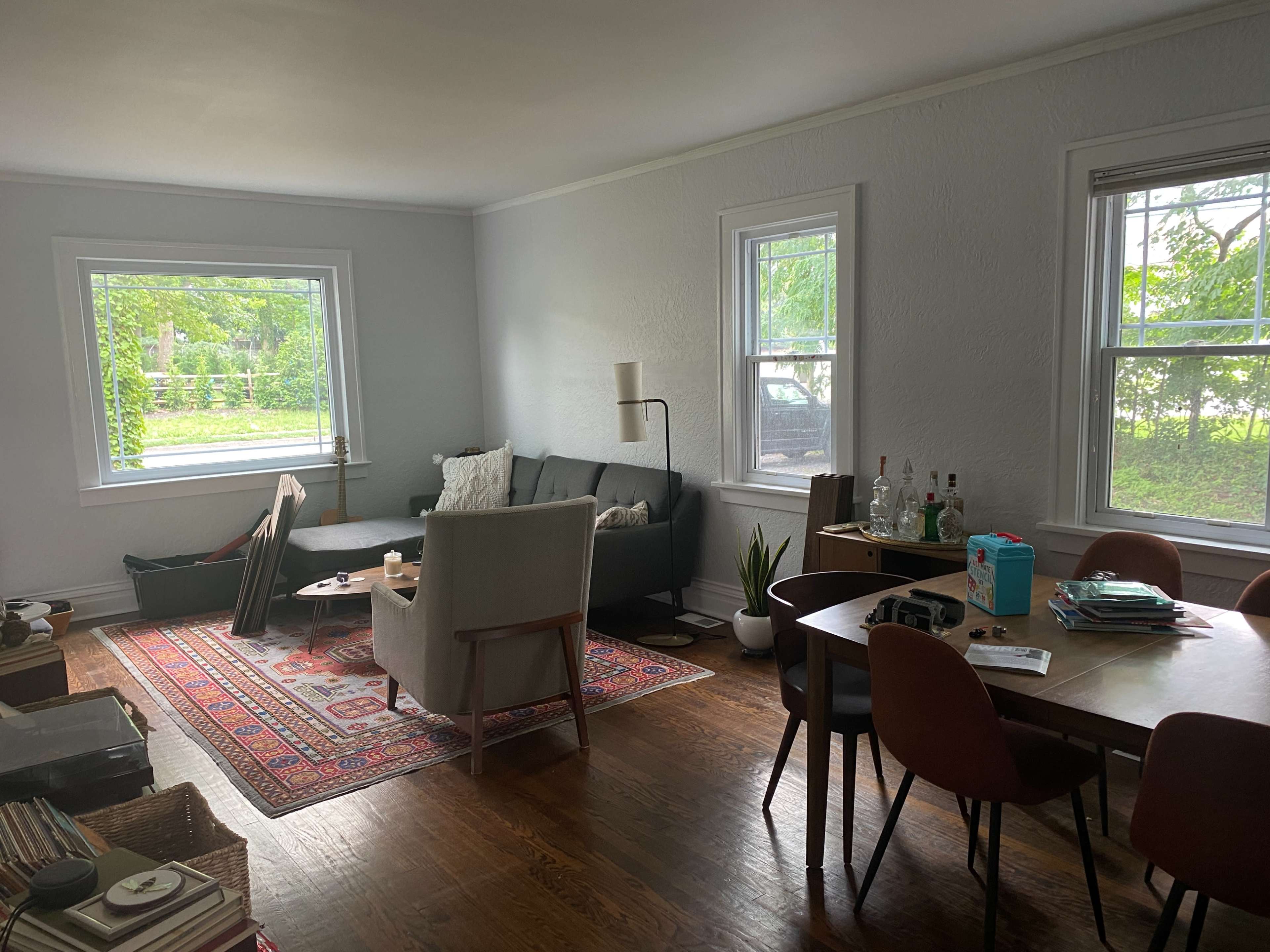 The living area features a gray sofa, a coffee table, and a dining table surrounded by chairs, with large windows illuminating the space.