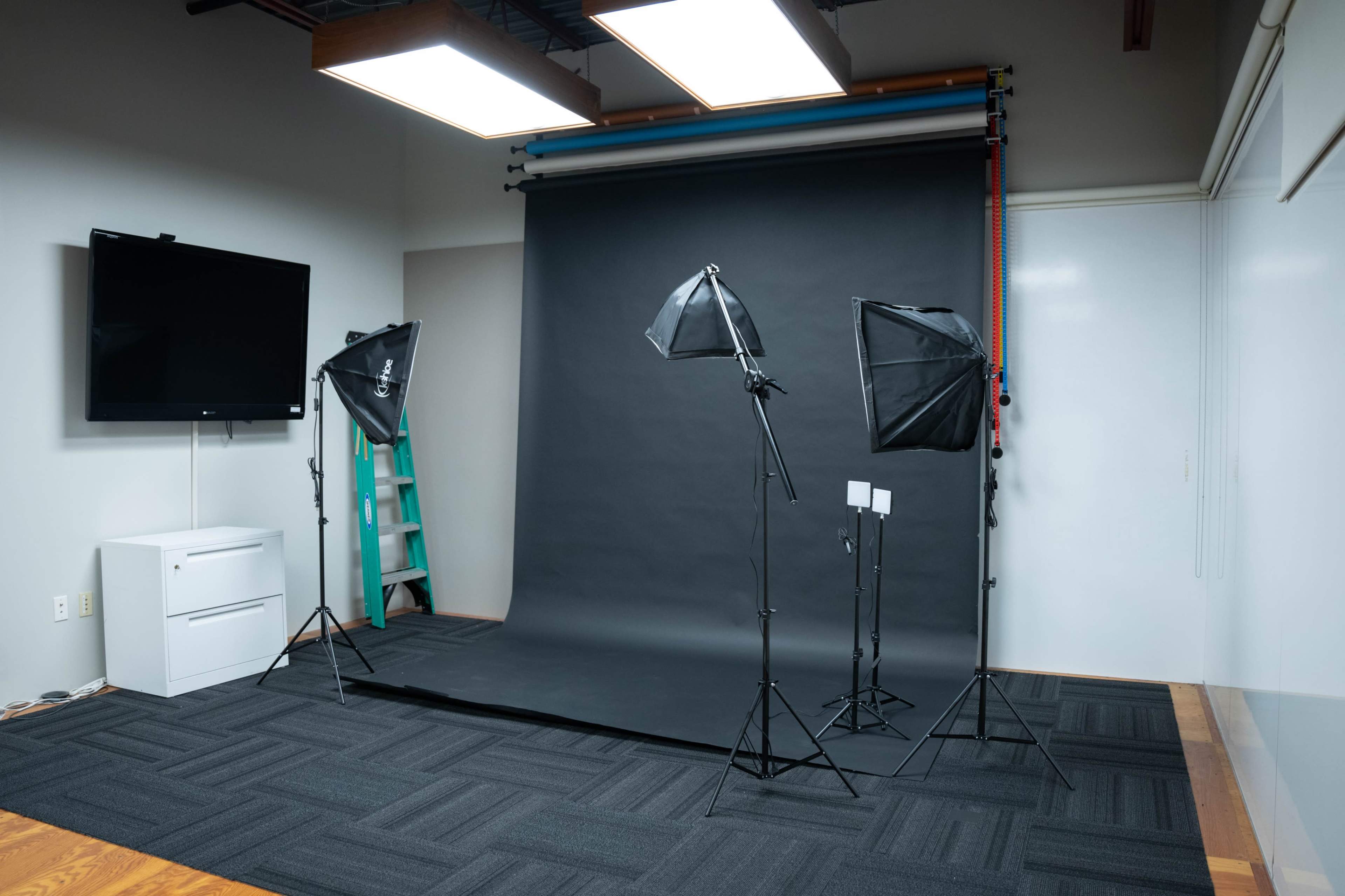 The image shows a photography studio equipped with a black backdrop, two softbox lights, a ladder, and a television on the wall.