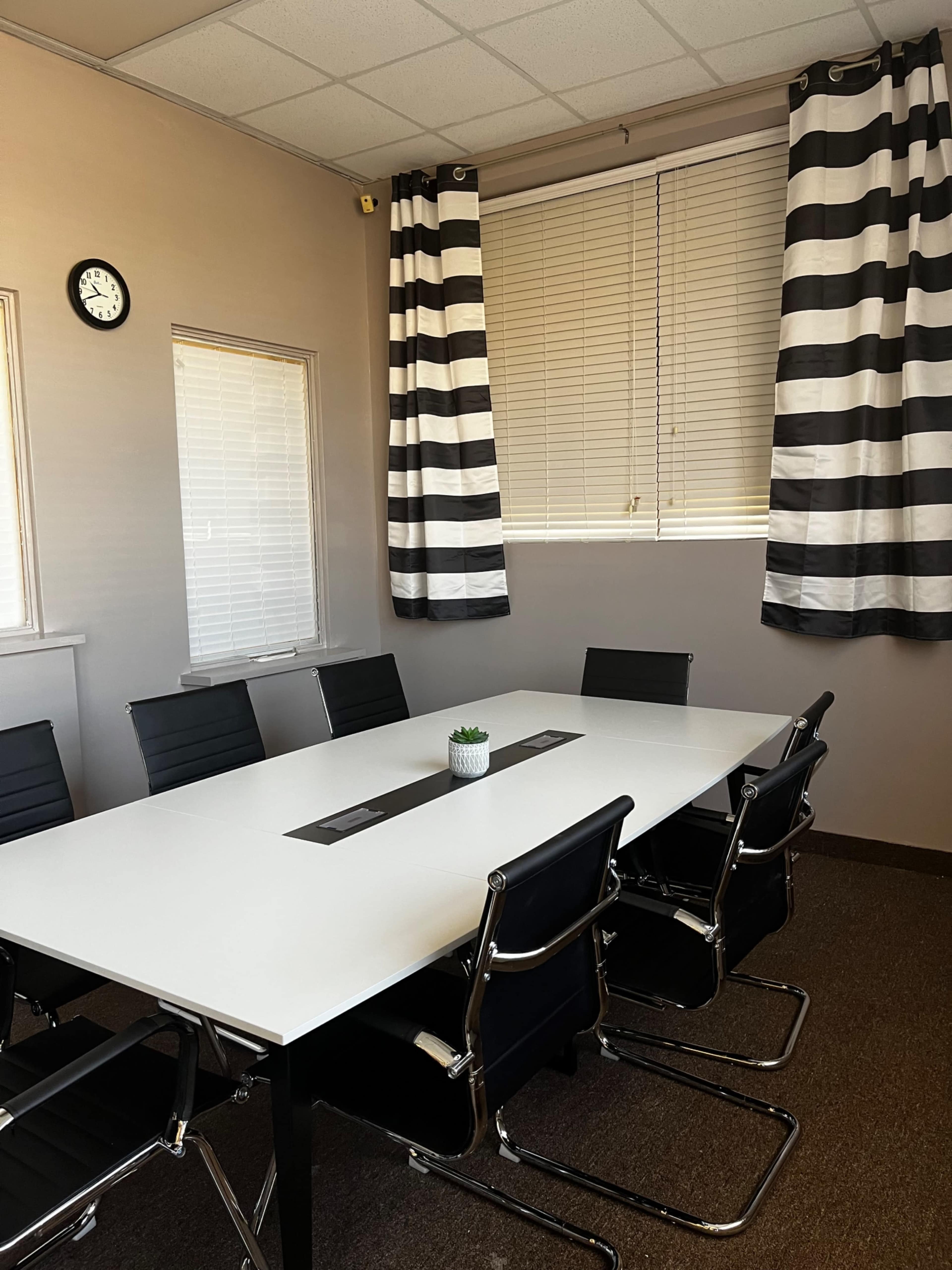 A conference room features a rectangular white table surrounded by black chairs, with striped curtains and a small plant on the table.
