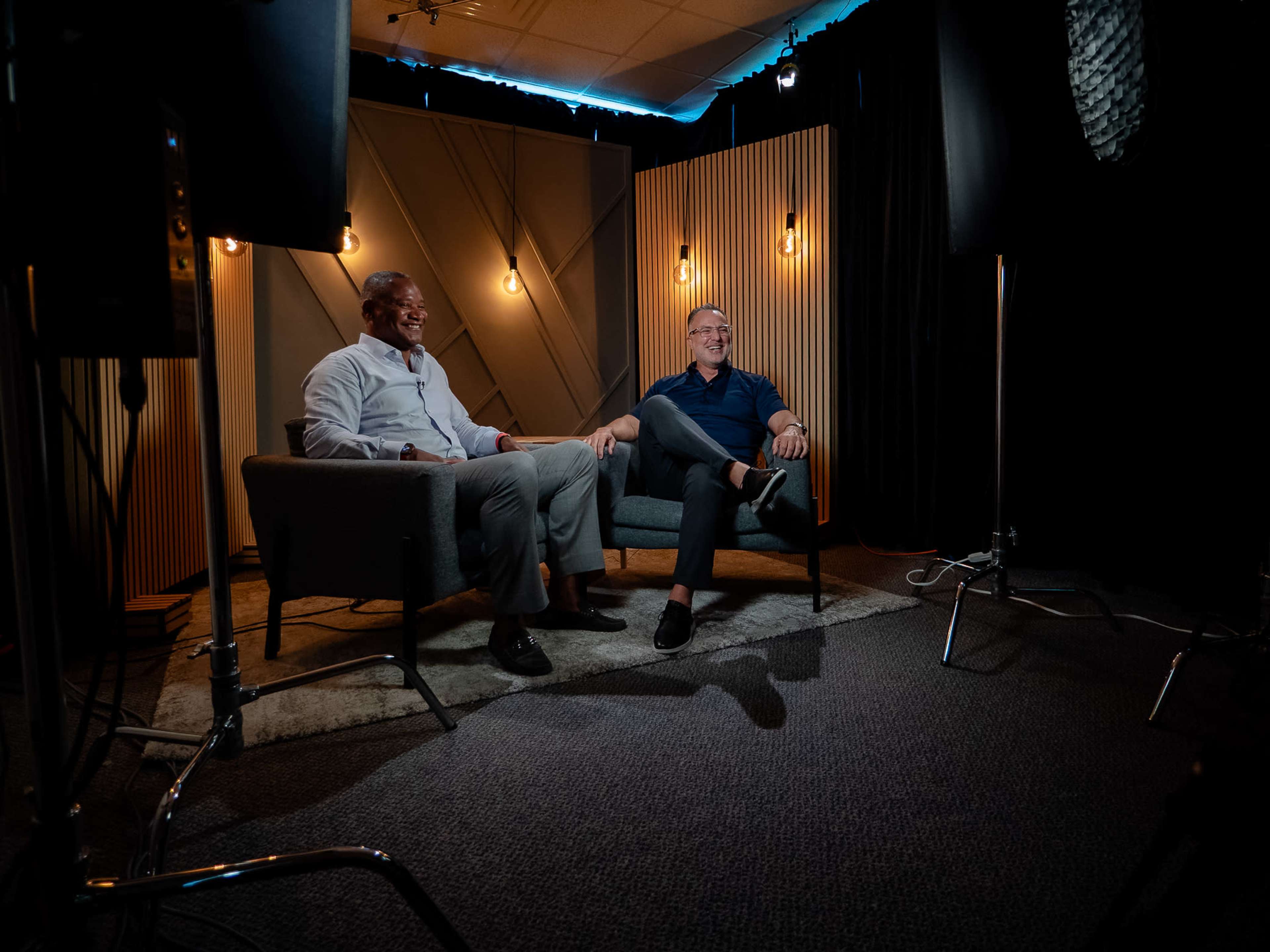 Two men sit on a gray couch in a dimly lit room with wooden paneling and light bulbs hanging in the background.