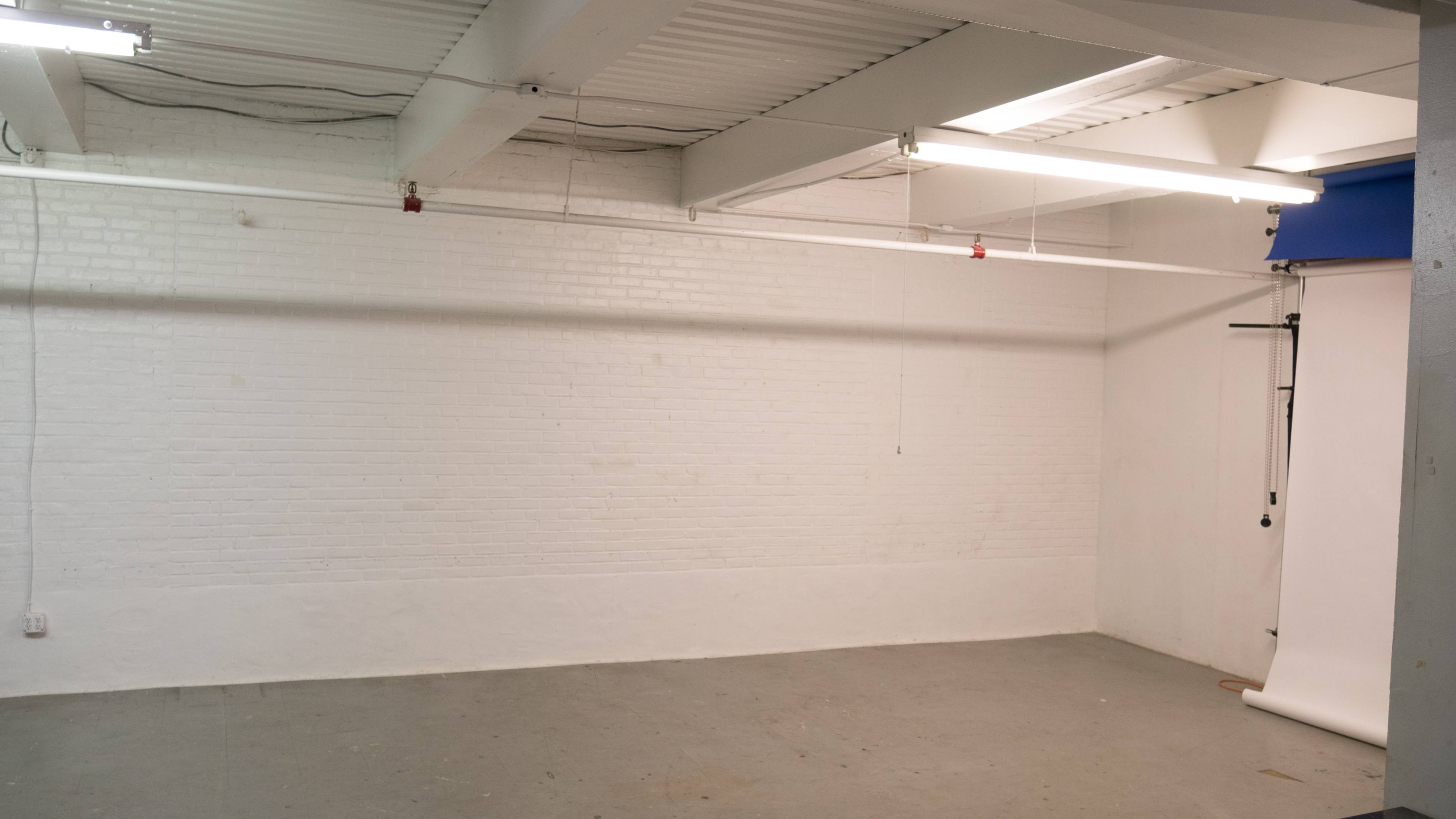 A minimally furnished photography studio with white brick walls and a gray floor, featuring overhead lighting and a blue backdrop.