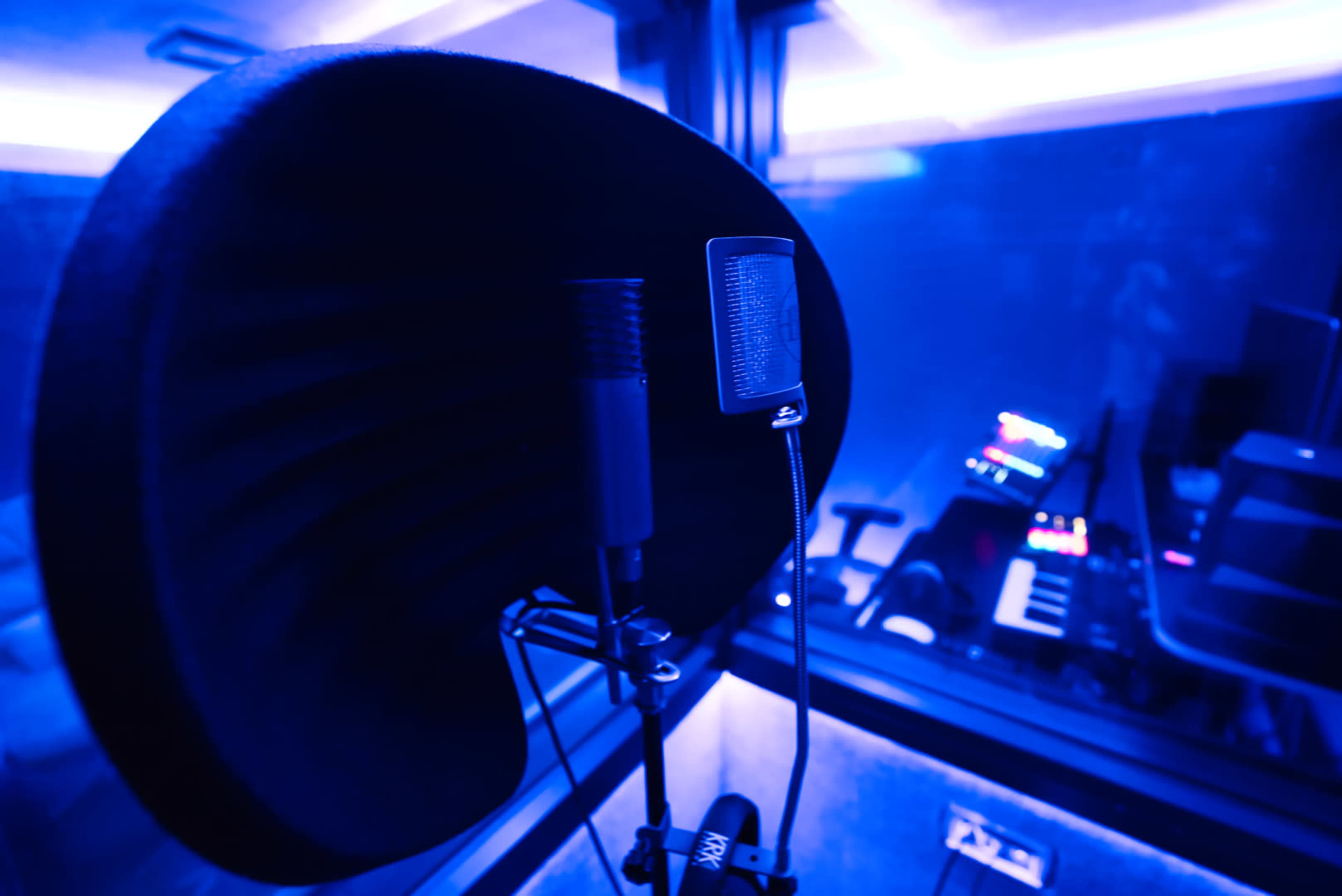 A close-up of two microphones positioned in a sound booth with blue lighting and a music keyboard visible in the background.