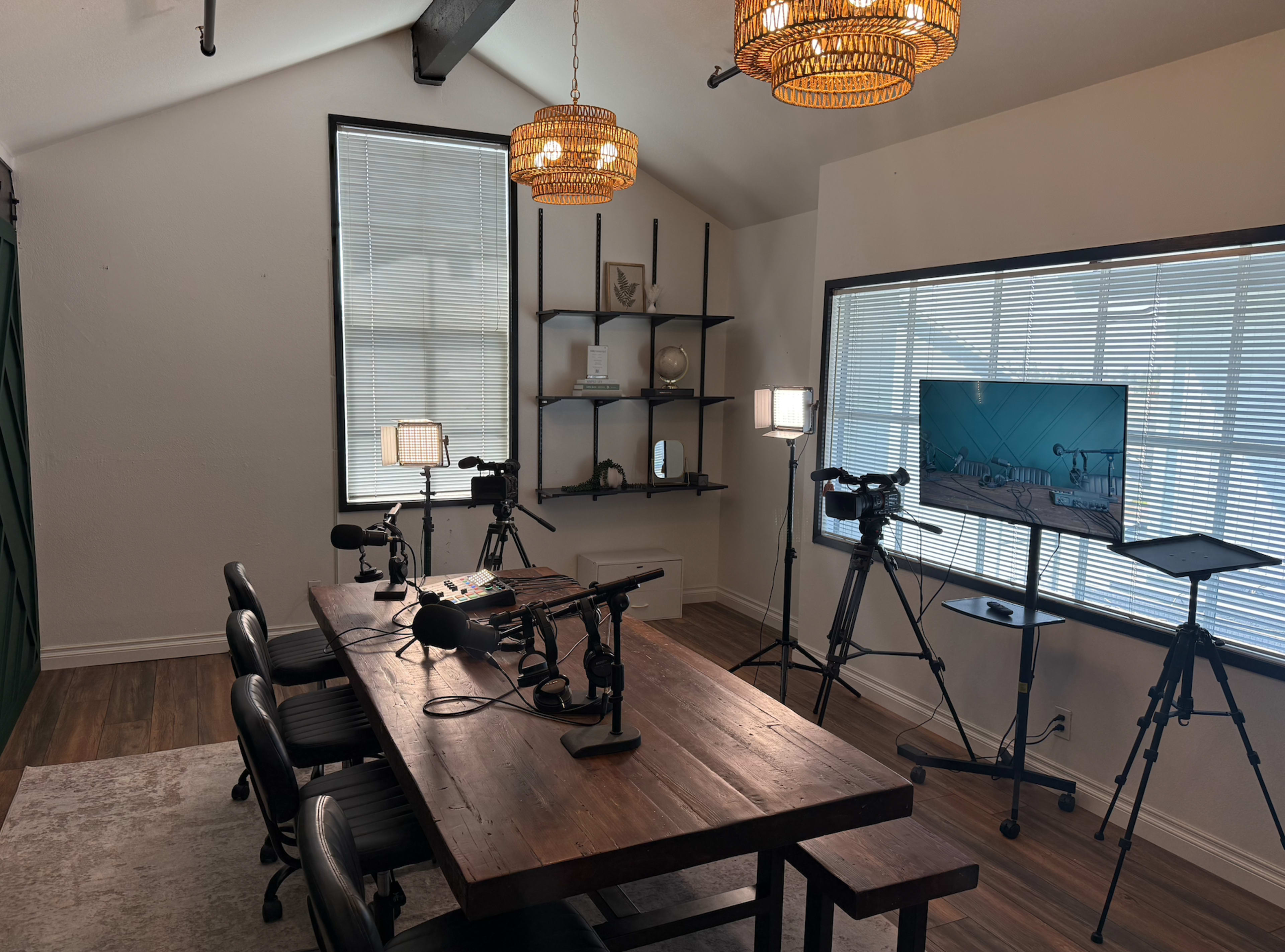 The image shows a modern room set up for a podcast or video recording, featuring a large wooden table with microphones and chairs, and equipment on tripods nearby.