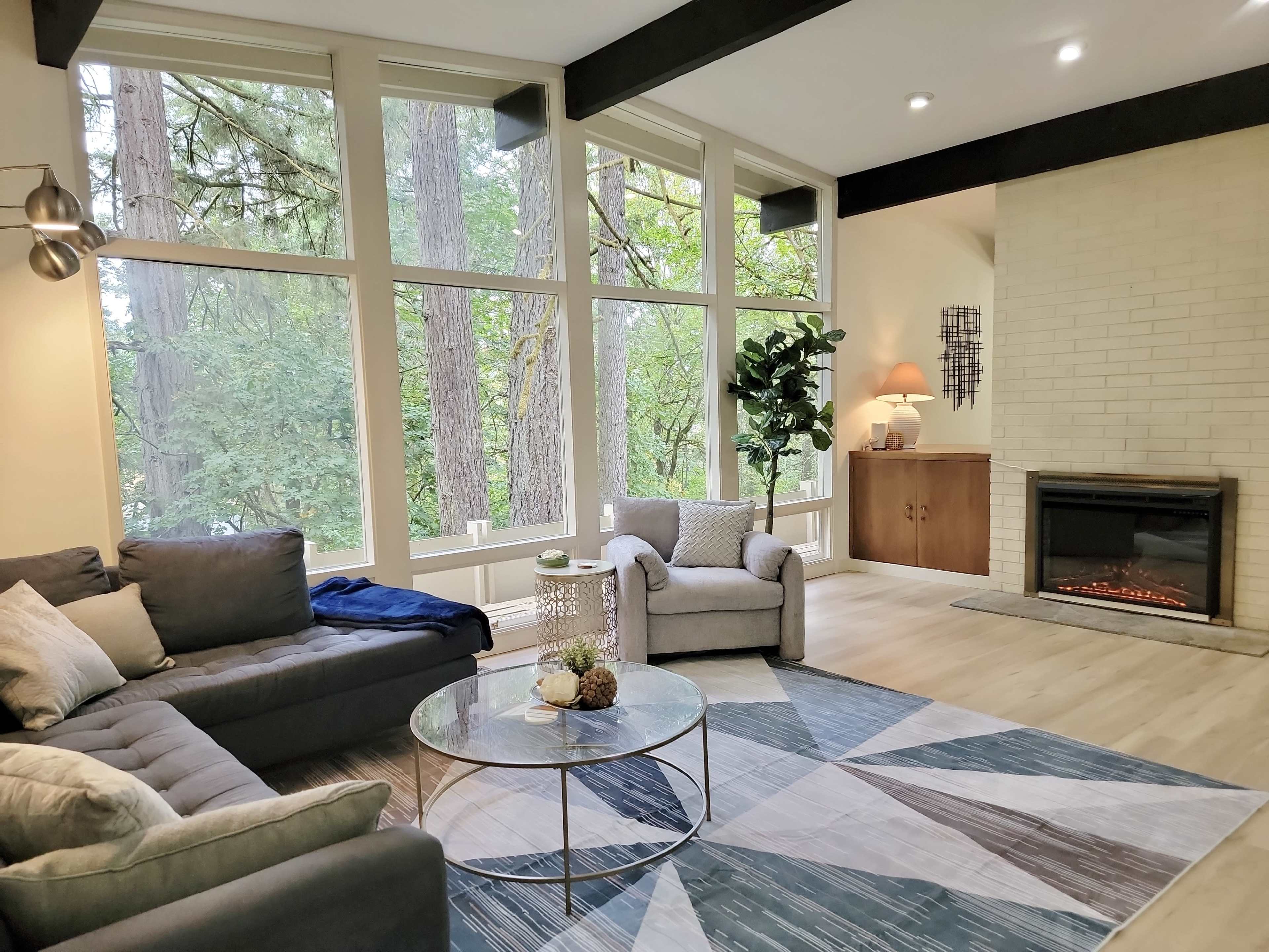 The image shows a modern living room with large windows overlooking a forest, featuring a gray sofa, a round coffee table, an armchair, and a fireplace.