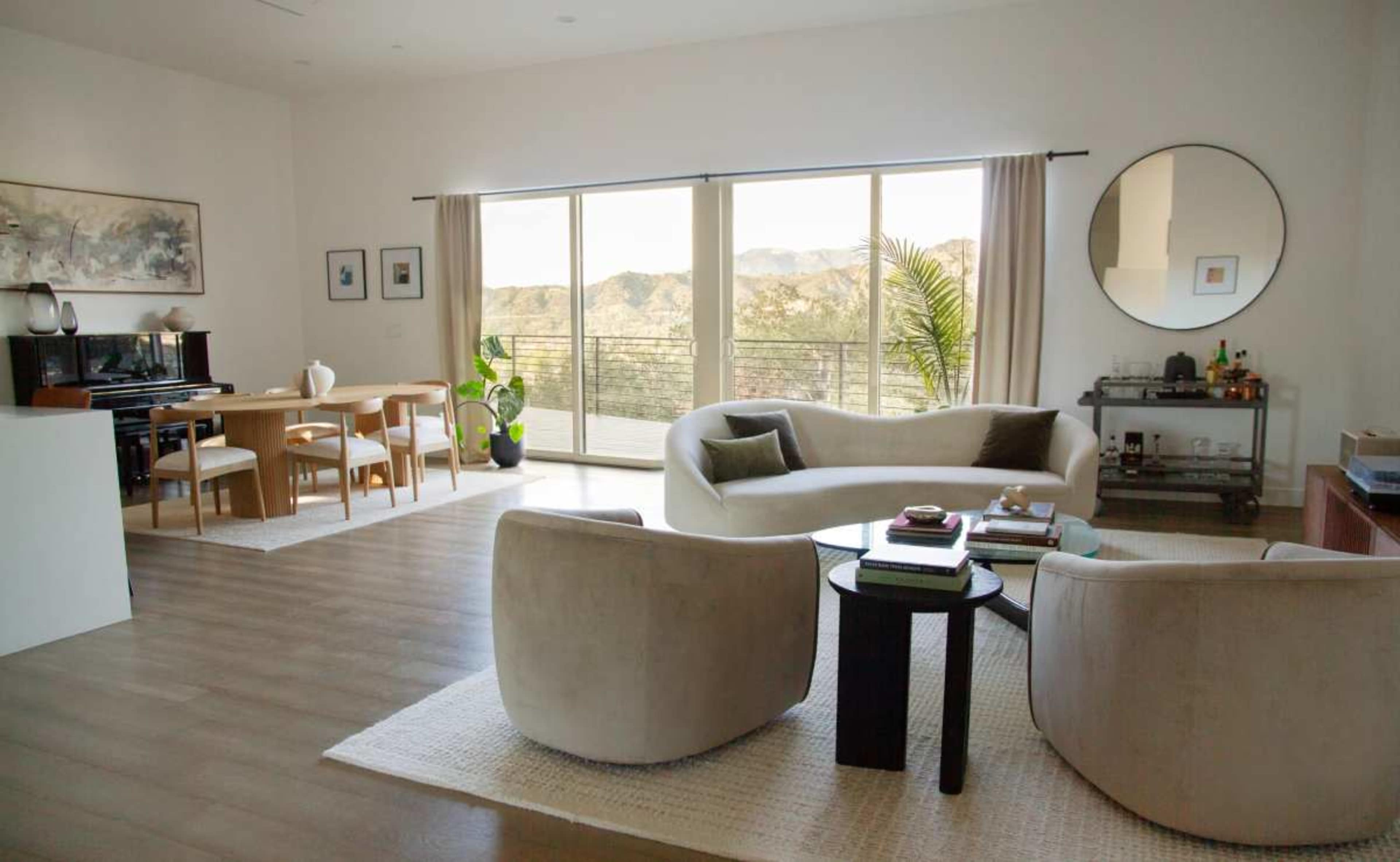 A modern living room features a curved sofa, two armchairs, a dining table, and large windows that overlook a mountainous landscape.