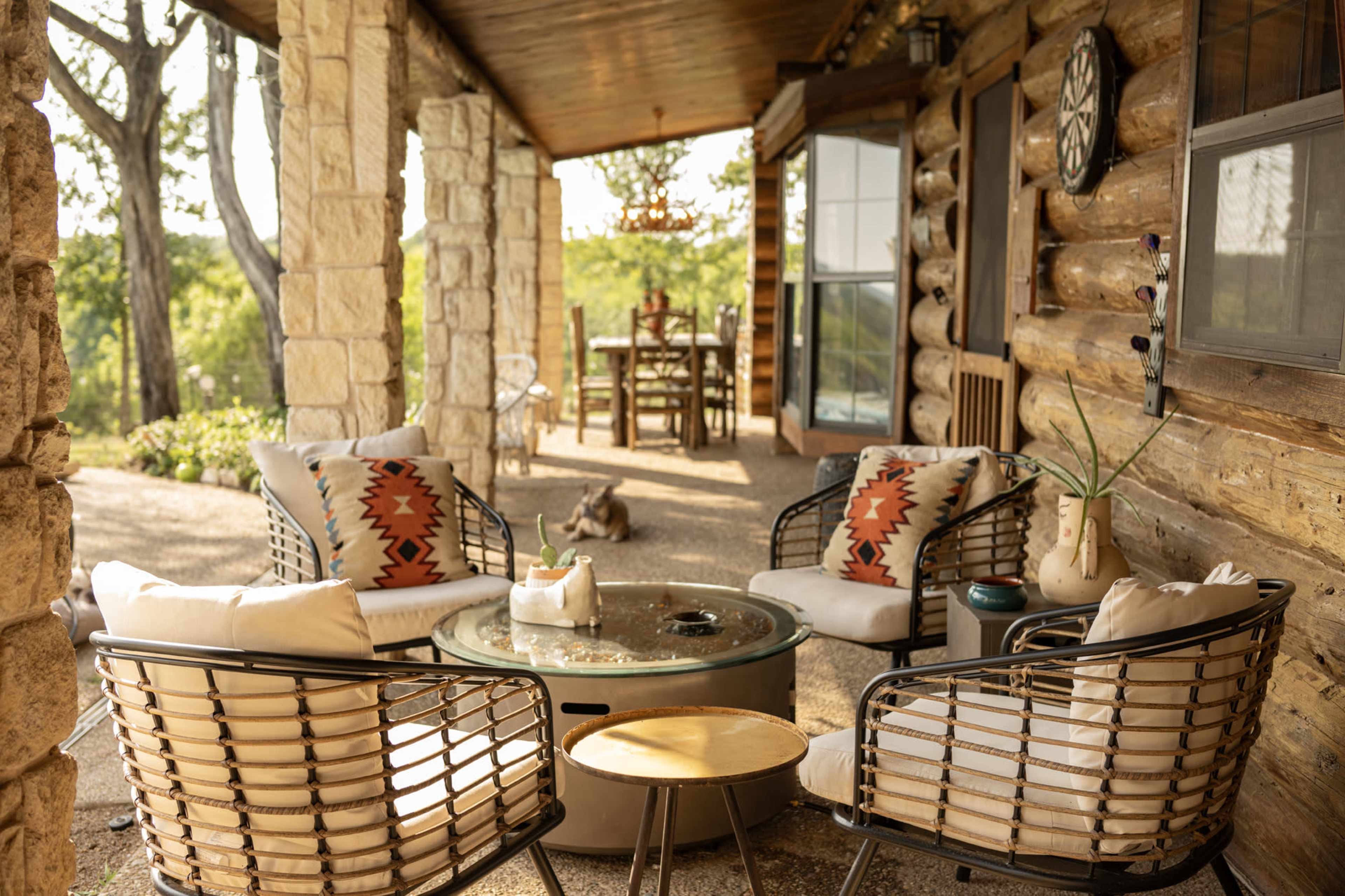 A rustic outdoor seating area with wicker chairs and a circular coffee table, set against a backdrop of a cabin porch and a view of green hills in the distance.