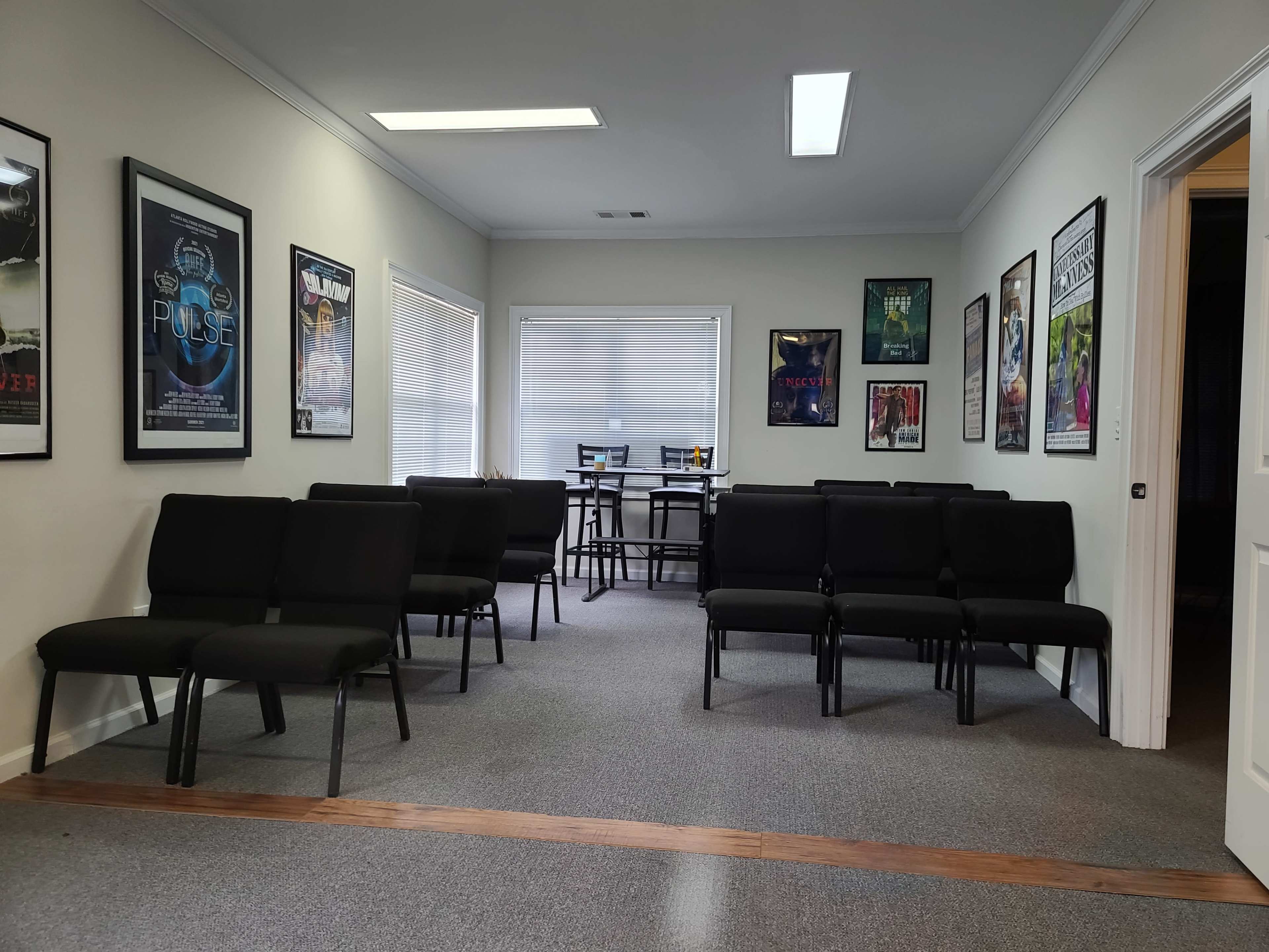A waiting room with rows of black chairs and movie posters on the walls, leading to a small table with chairs in the back.