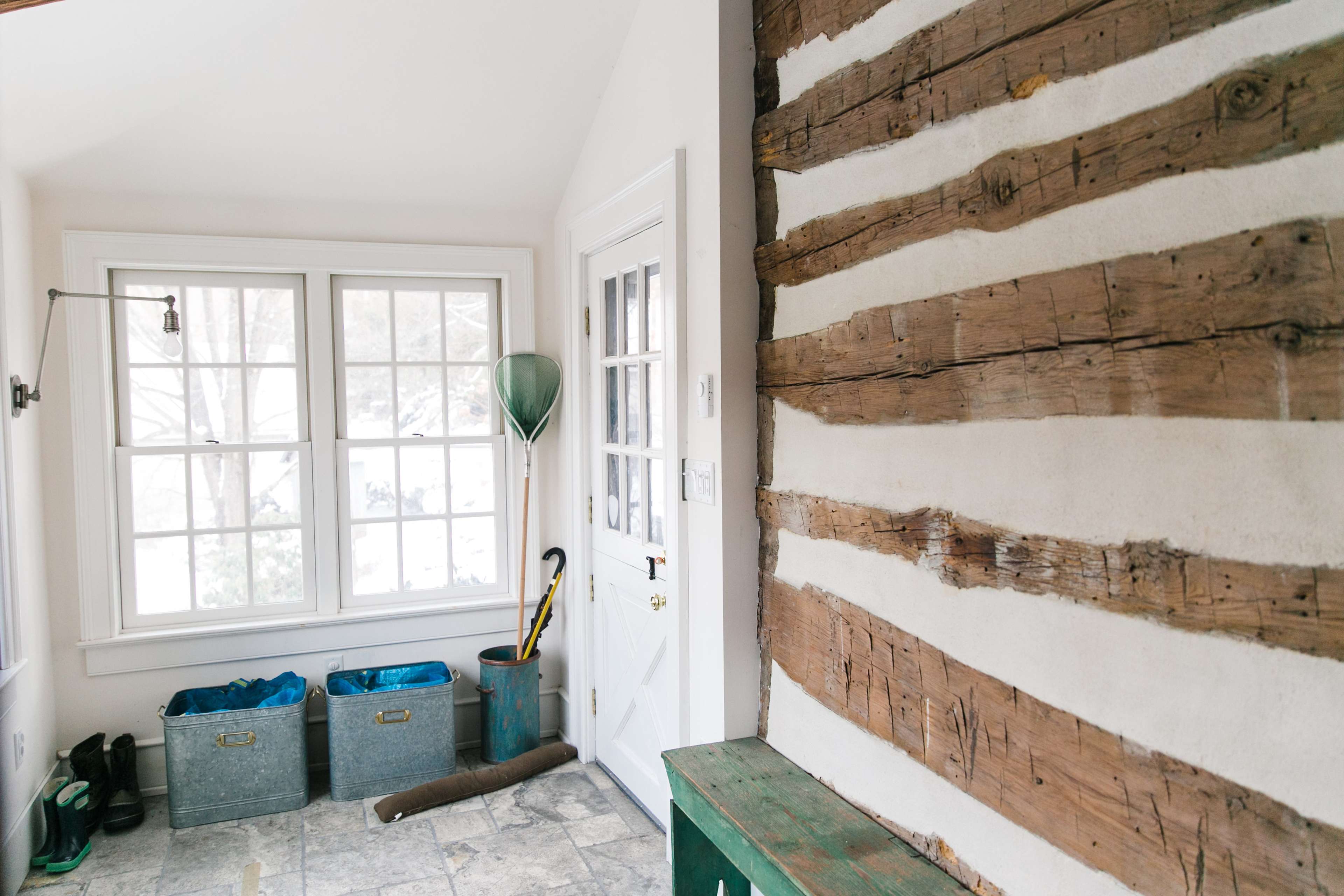 An interior space featuring a rustic wooden wall, large windows allowing natural light, storage bins, and a door with an umbrella nearby.
