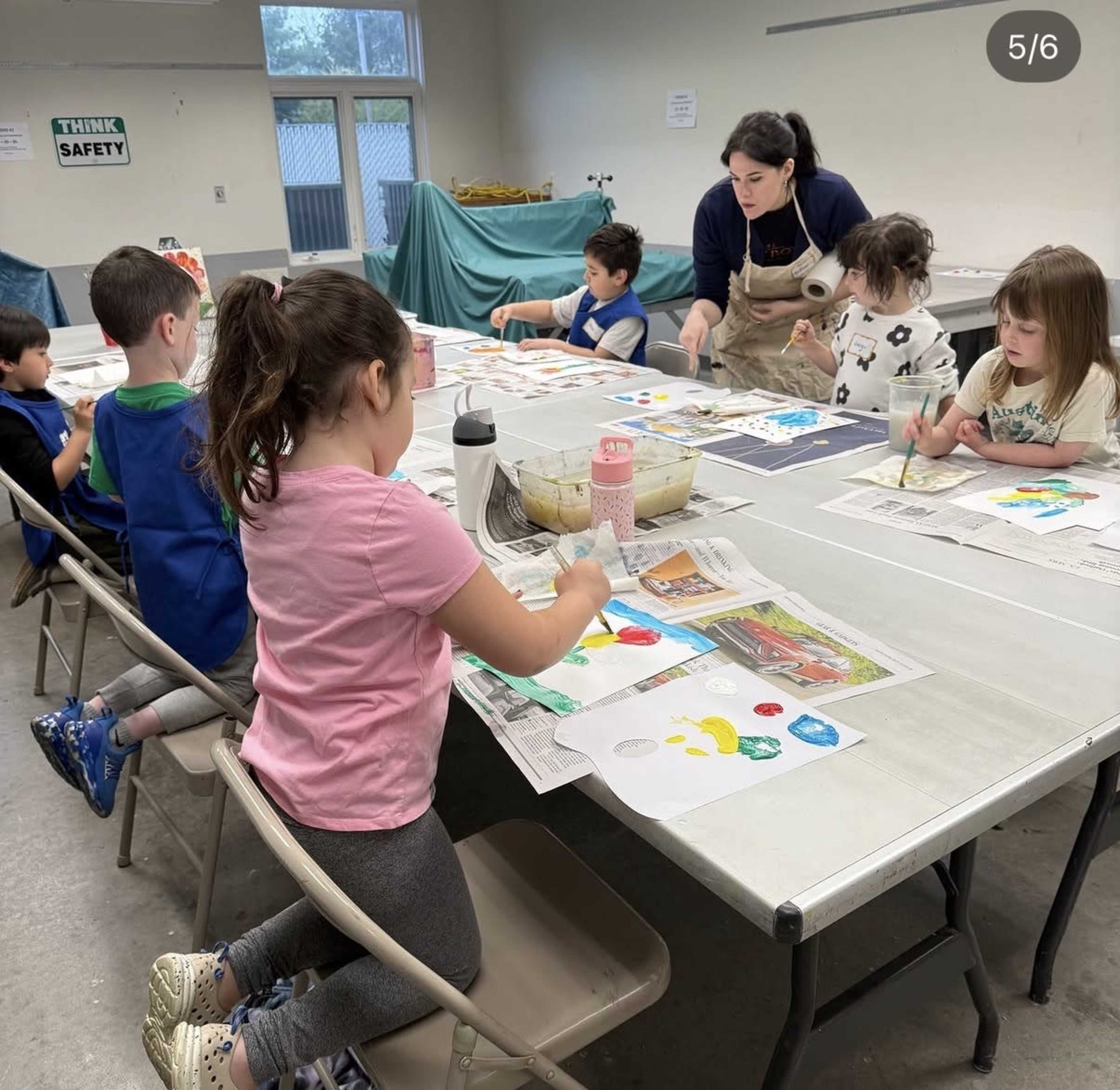 A group of children is painting at a long table while a woman gives guidance in an indoor classroom setting.