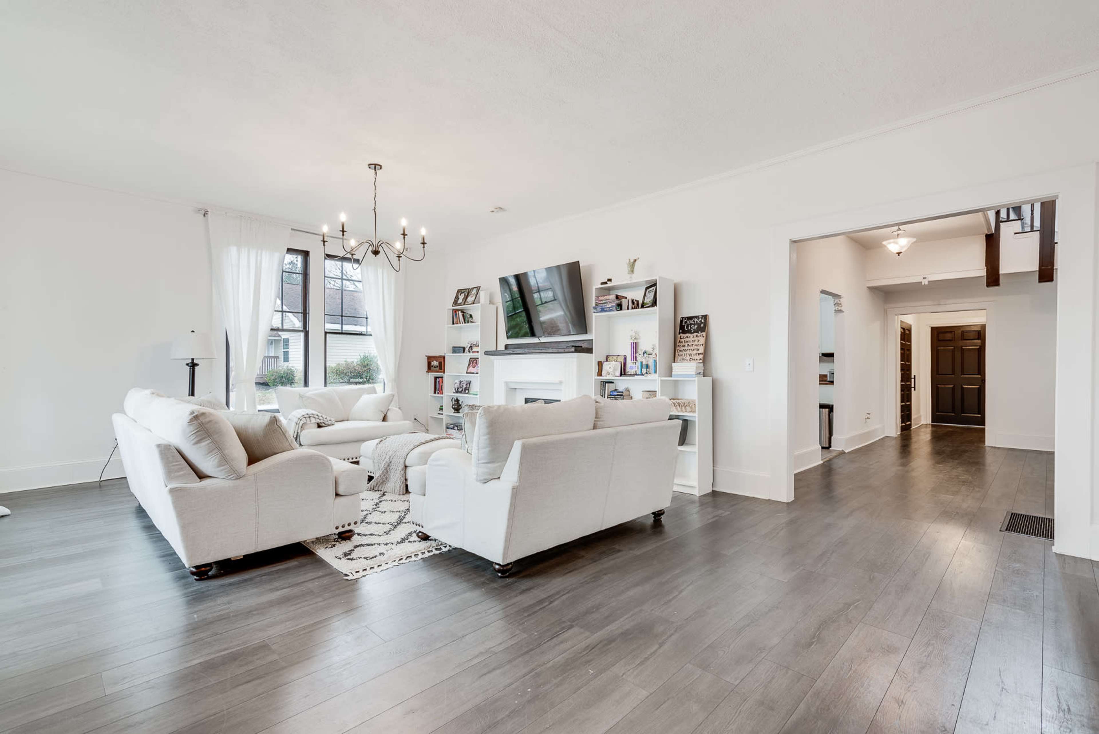 A spacious, modern living room with two white sofas, a large television mounted on the wall, and an entrance leading to a hallway.