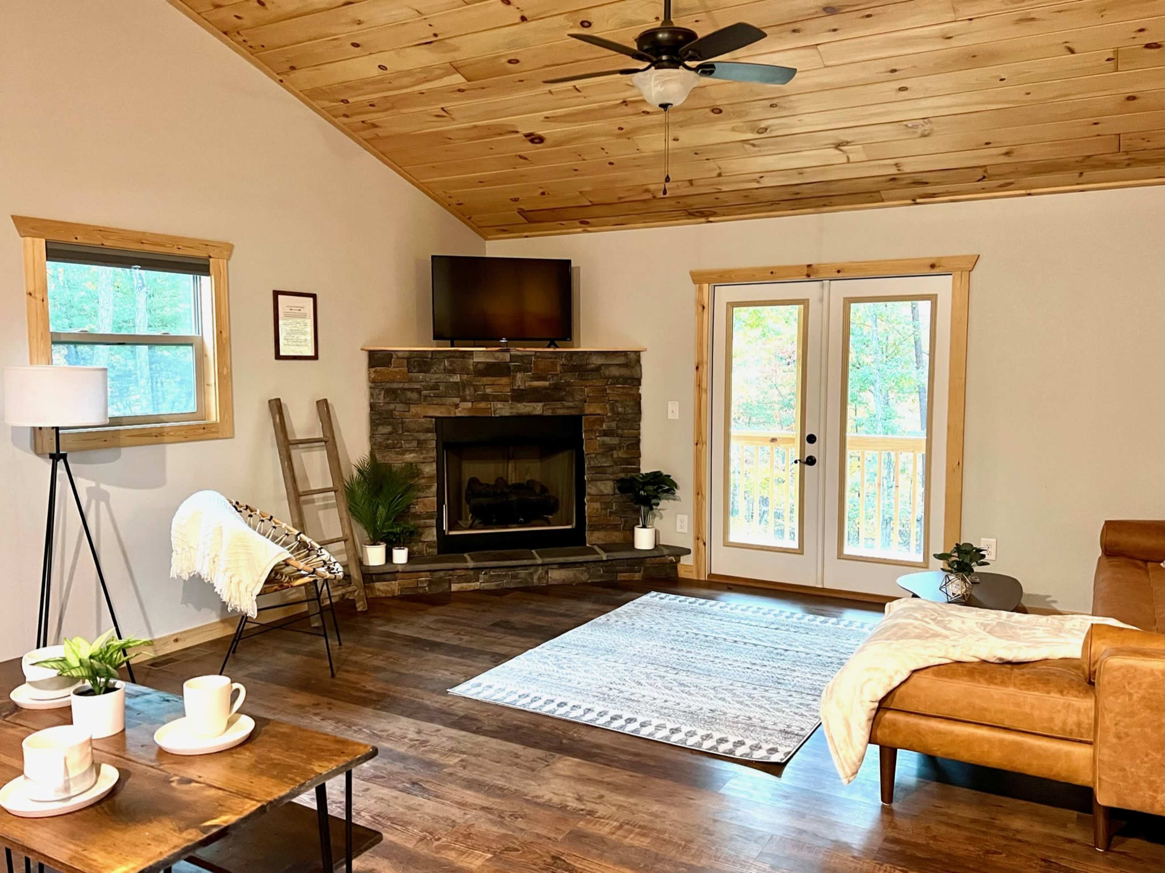 The image shows a cozy living room with a stone fireplace, a wooden ceiling, a brown leather sofa, and a large window with a door leading outside.