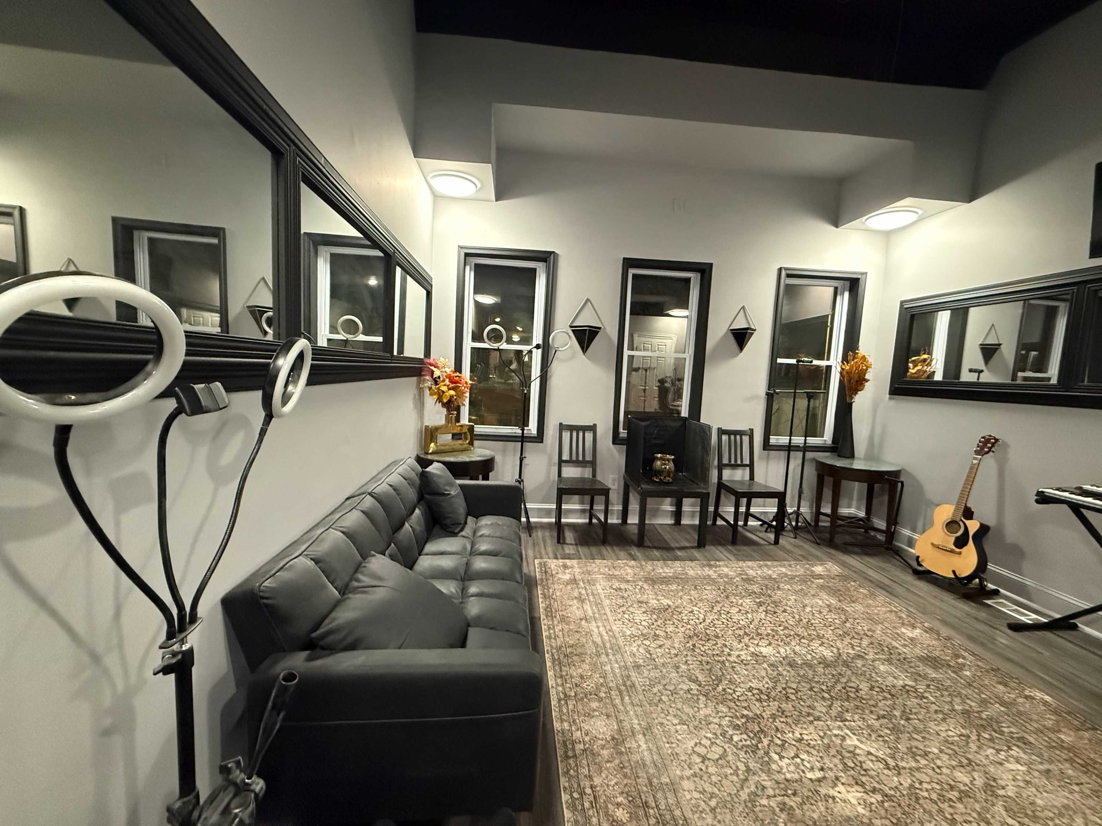 The image shows a modern room with a black sofa, several mirrors, two chairs, a guitar, and decorative elements in a well-lit space.