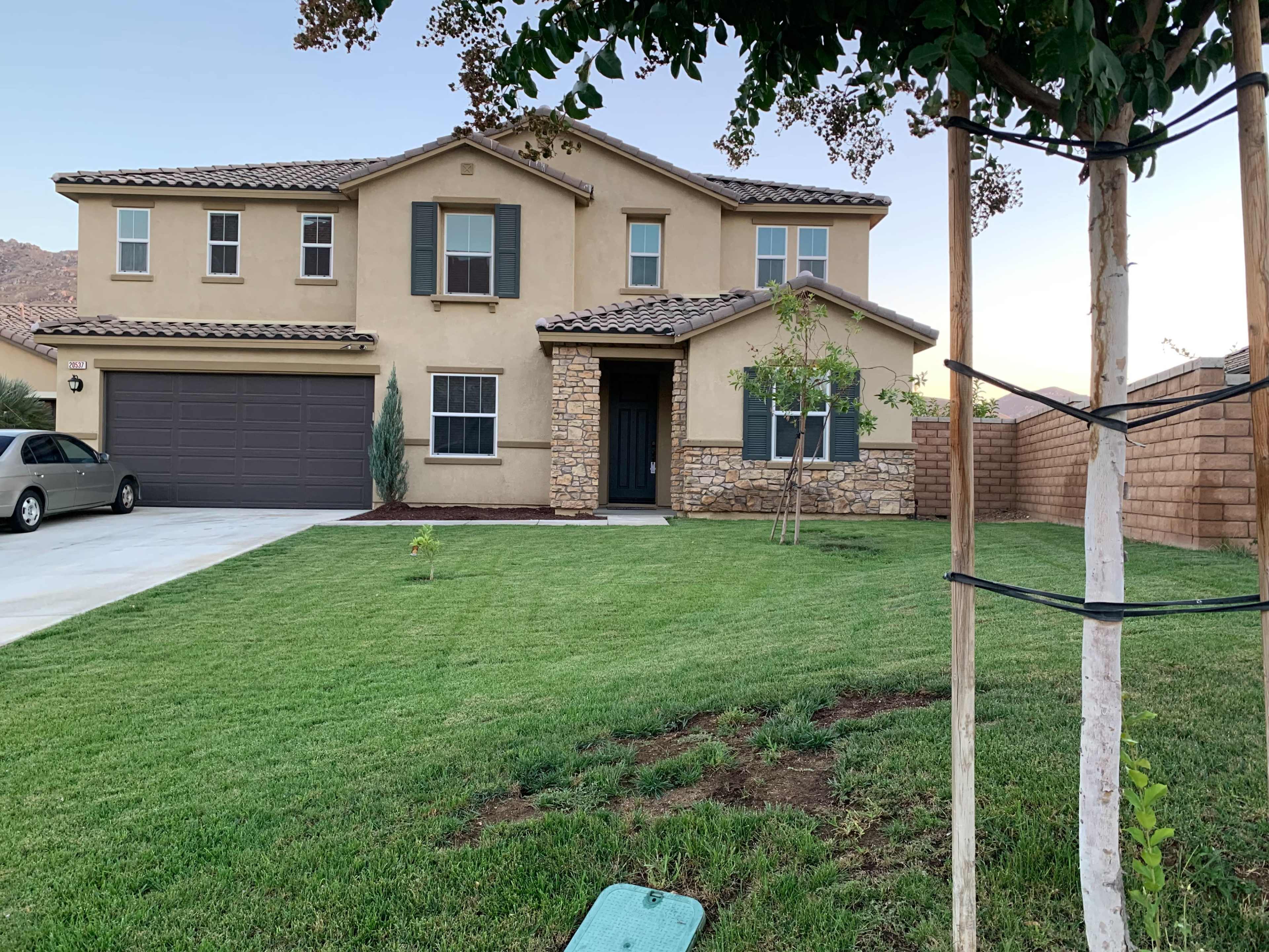 A two-story house with a stone accent, a green lawn, and a driveway, surrounded by partial fencing and a small young tree.