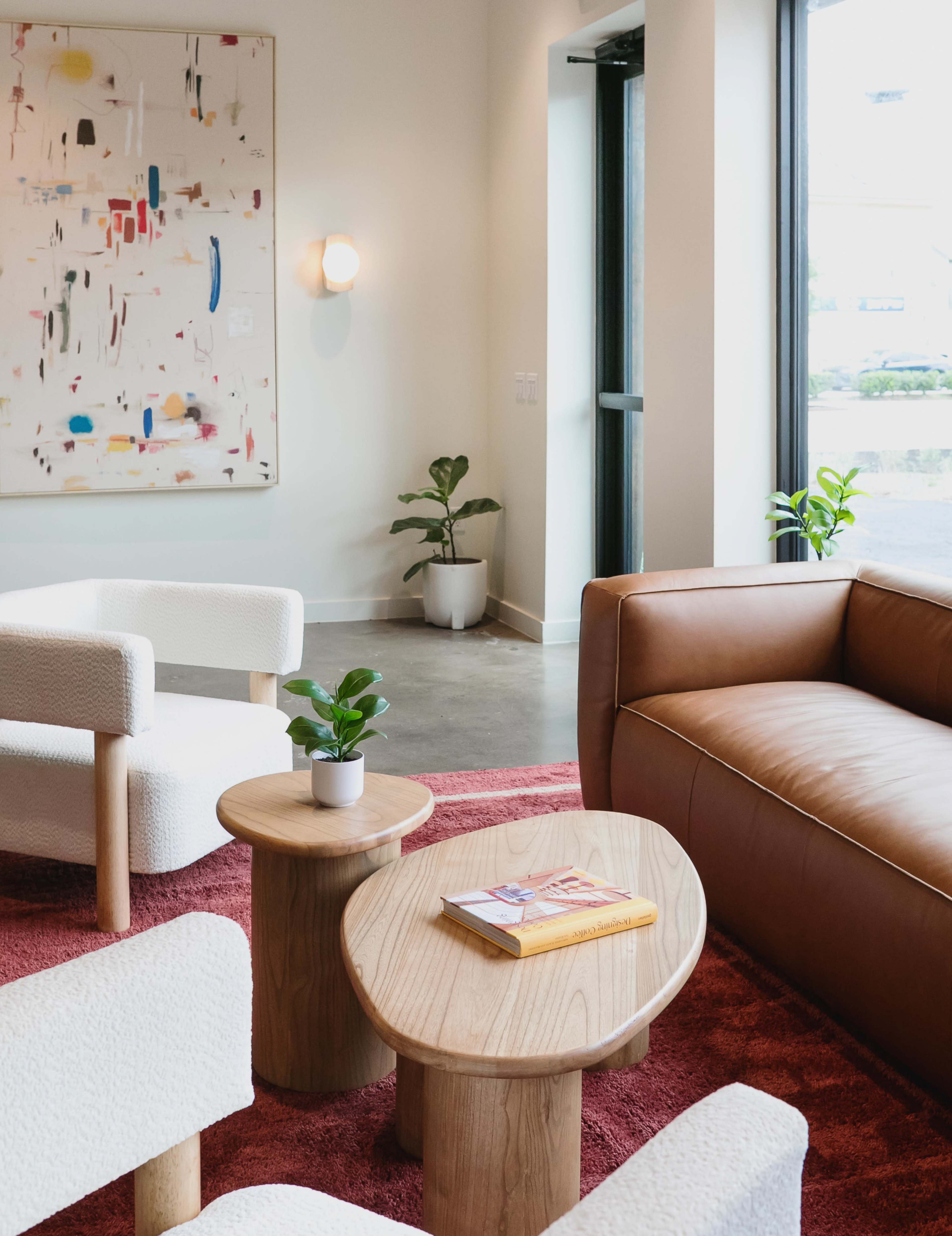A modern lounge area features a brown leather sofa, textured white chairs, a round wooden table, and a colorful abstract artwork on the wall.