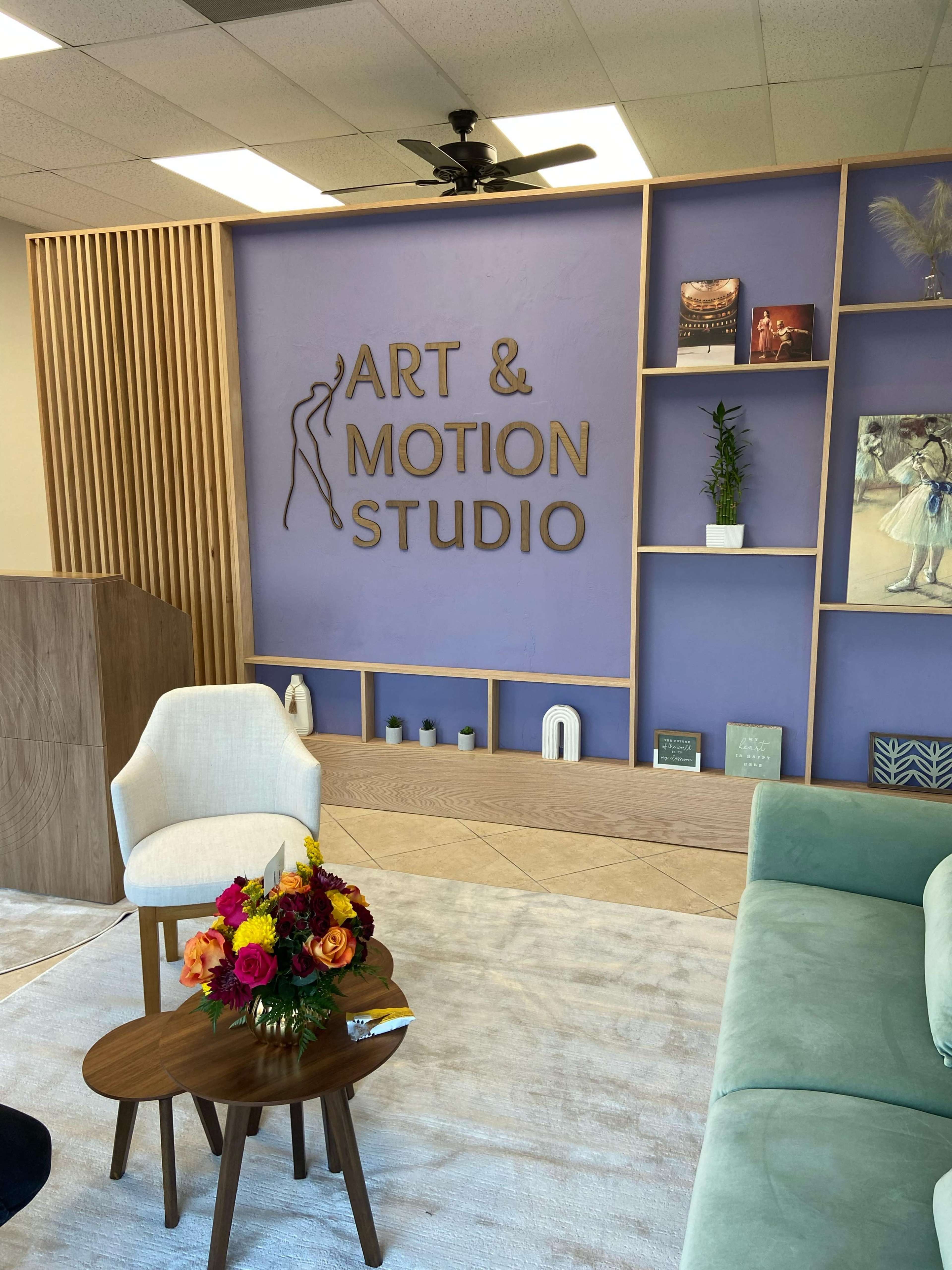 The image shows a modern studio space featuring a purple accent wall with the words "Art & Motion Studio" in gold lettering, decorated with plants and art, alongside a seating area with a round coffee table and a flower arrangement.