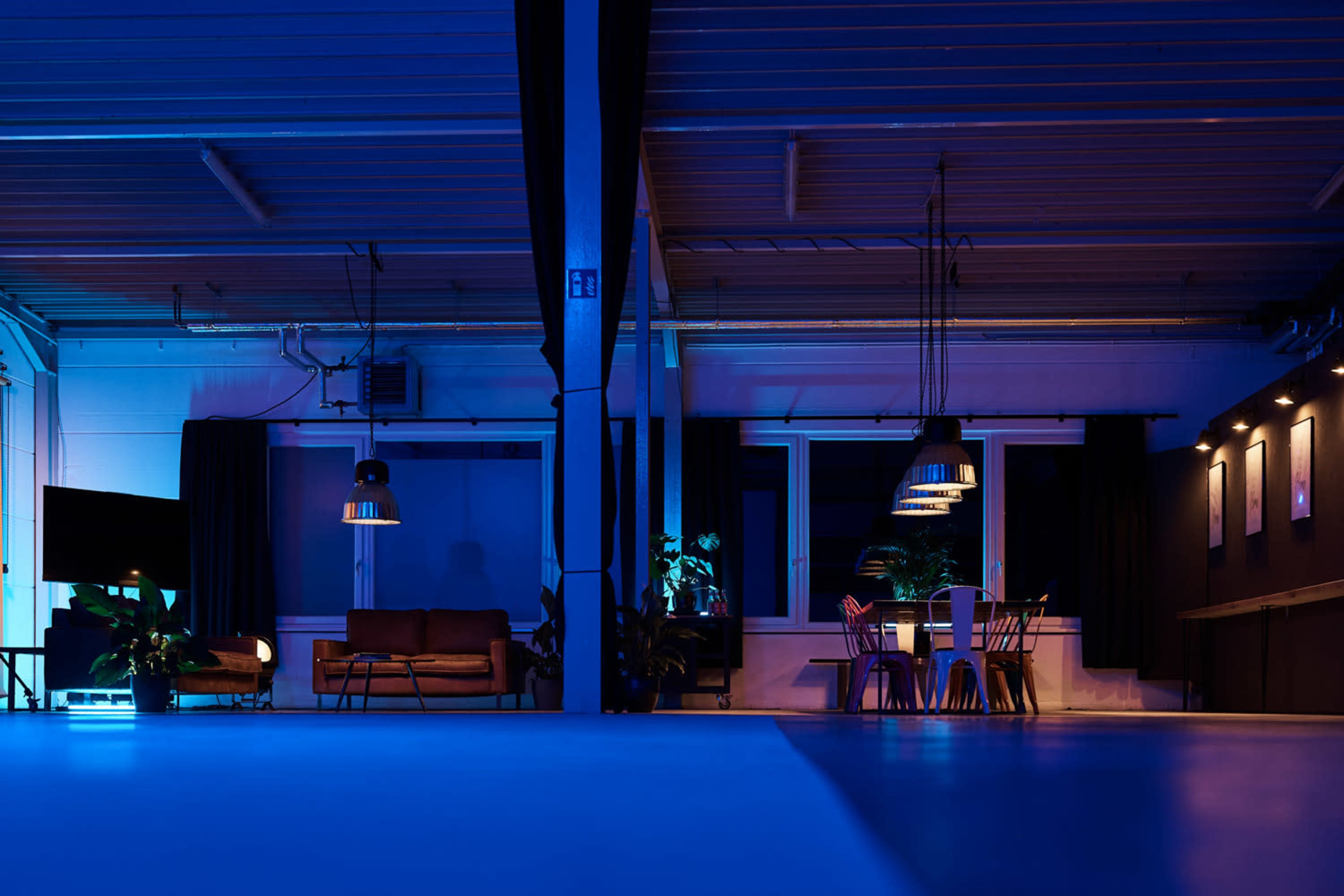 A spacious room with blue lighting, featuring a couch, dining table with chairs, and plants against a backdrop of large windows.