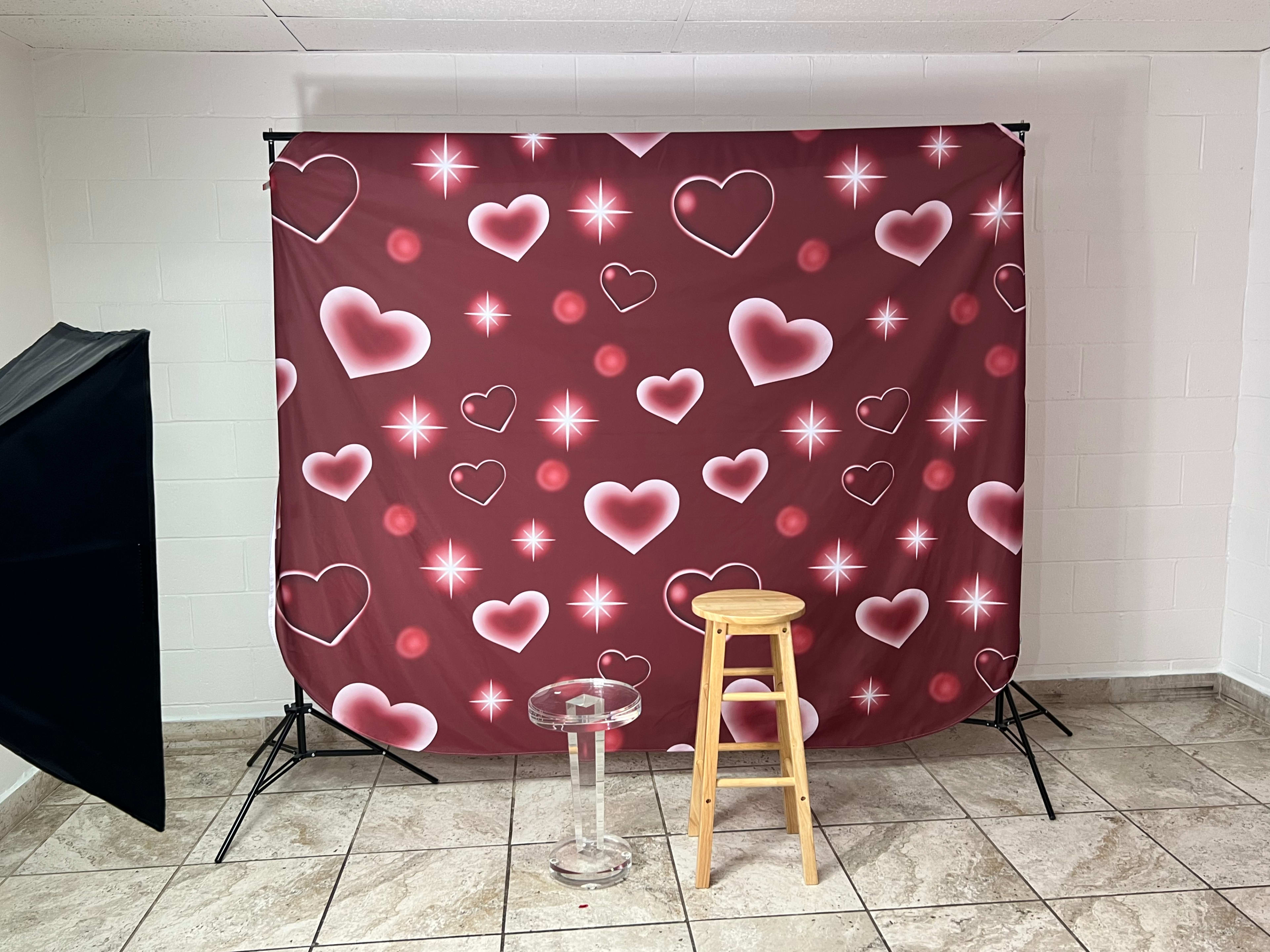A photography backdrop featuring a pattern of hearts and stars in shades of pink and red, set up in a room with a stool and a glass table in front of it.