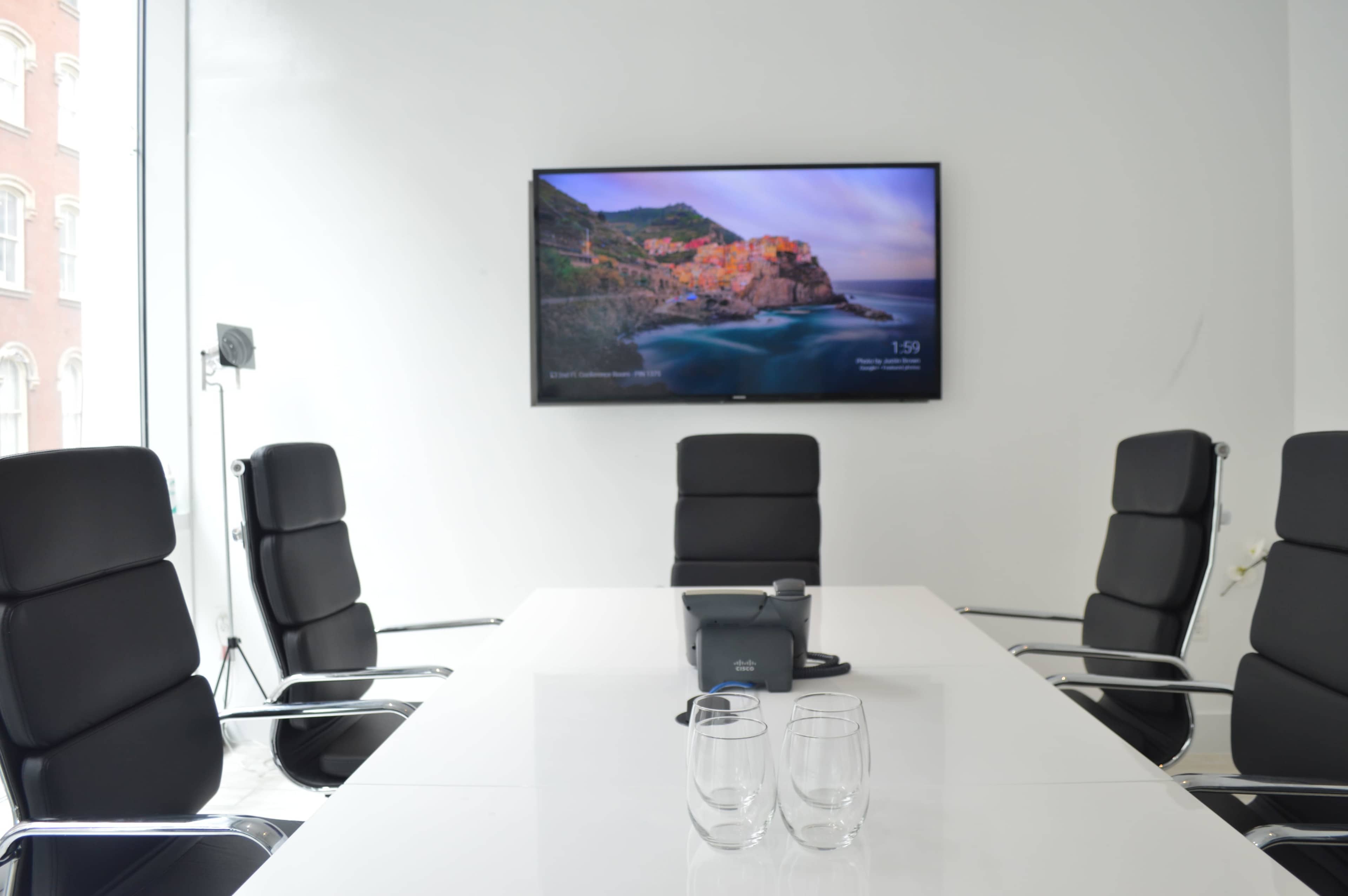 A modern conference room features a white table surrounded by black chairs, with a large screen displaying a coastal landscape on the wall.