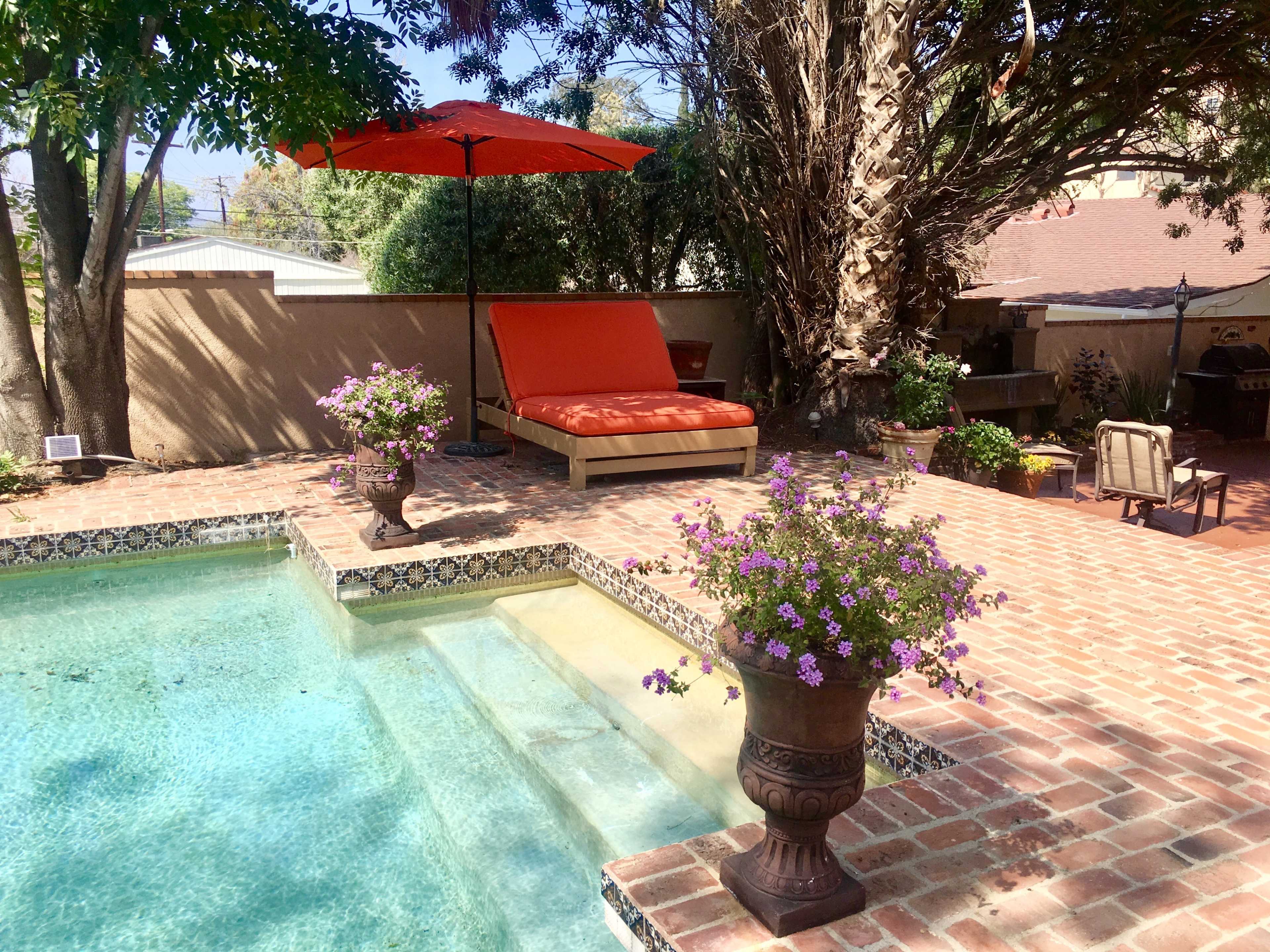A backyard pool area featuring a lounge chair under an orange umbrella, surrounded by potted flowers and brick patio.