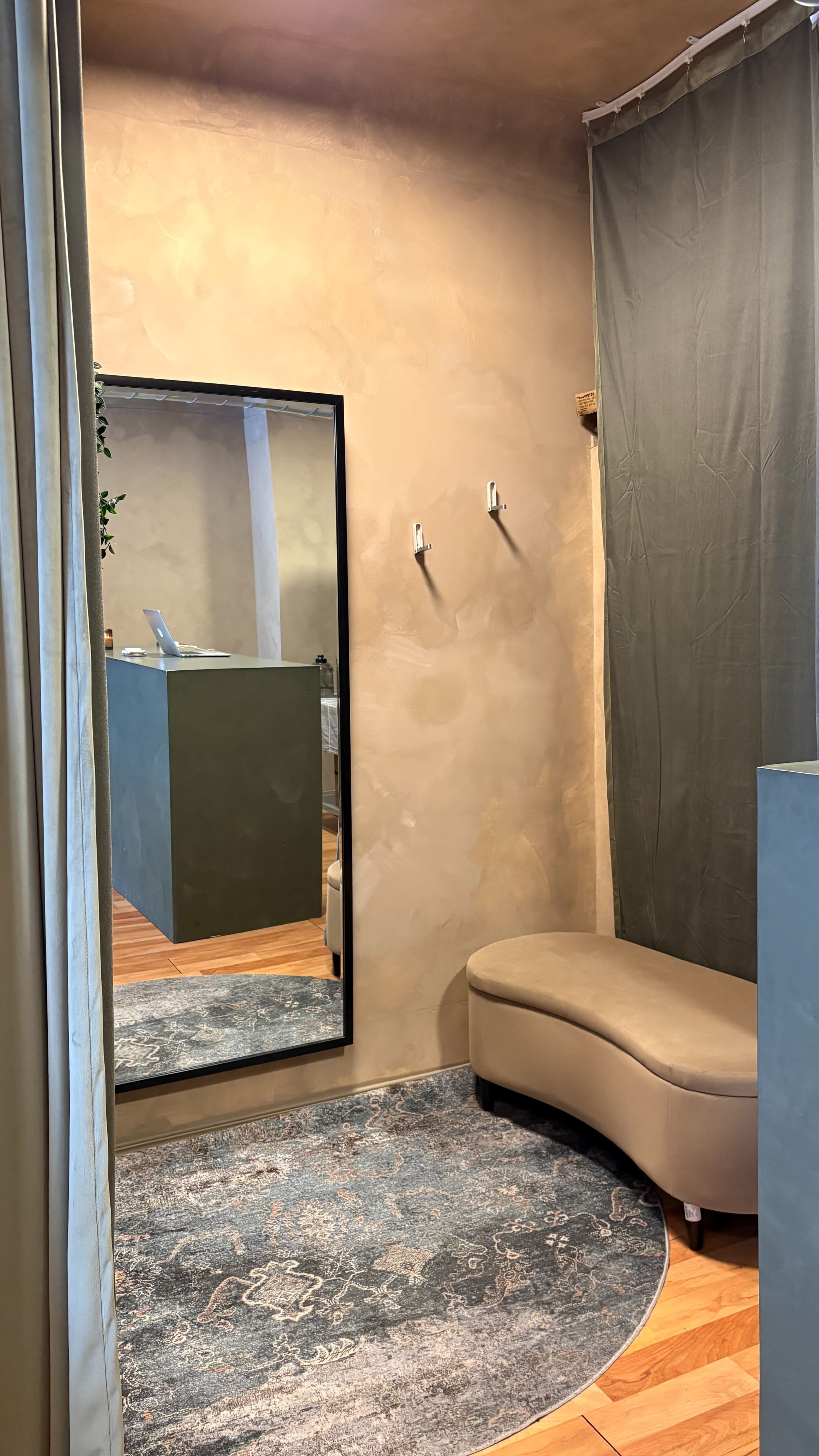 The image shows a small, minimalist dressing area featuring a curved bench, a full-length mirror, and a muted fabric curtain, with walls finished in a warm beige tone.