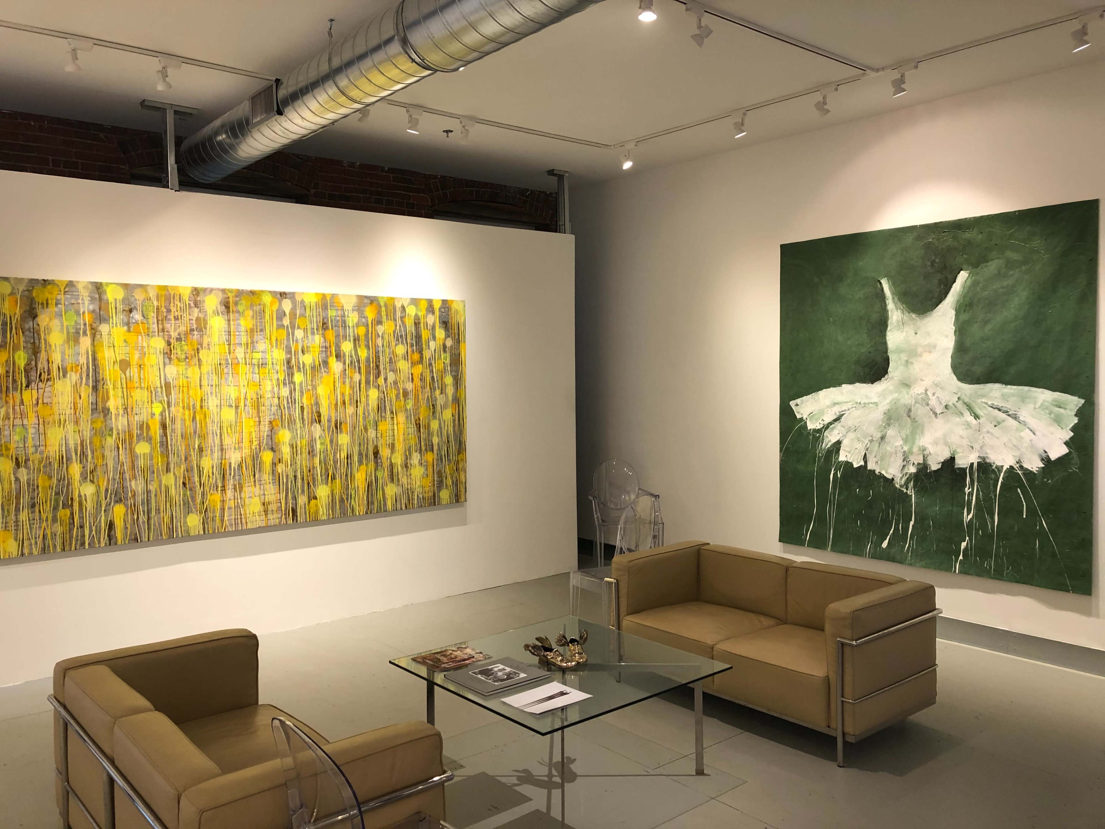 The image shows an art gallery with two large paintings on the walls and a seating area featuring two tan couches and a glass coffee table.