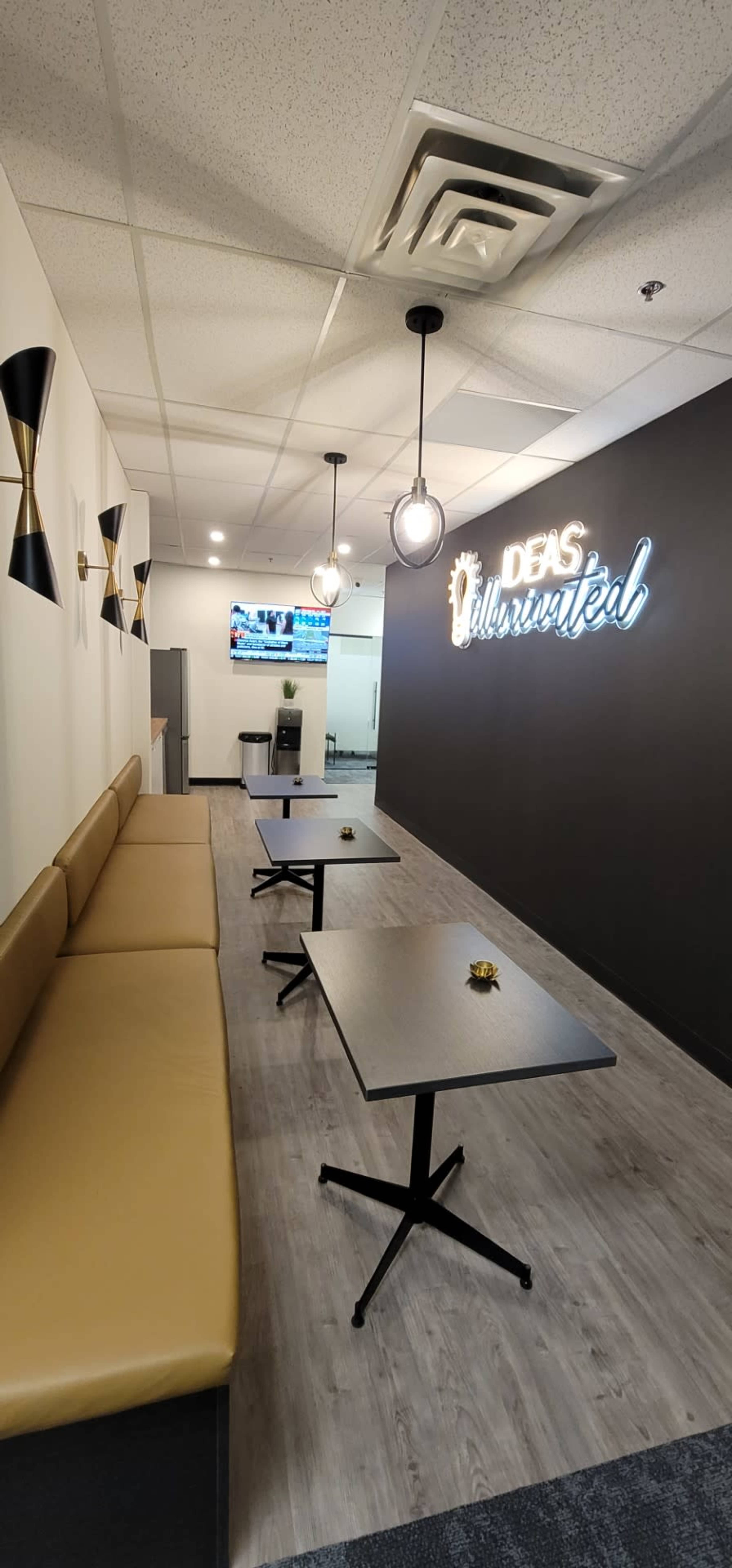 A modern workspace with a seating area featuring a tan bench and several black tables, illuminated by pendant lights, and a wall displaying the phrase "Ideas Unlimited" in neon.