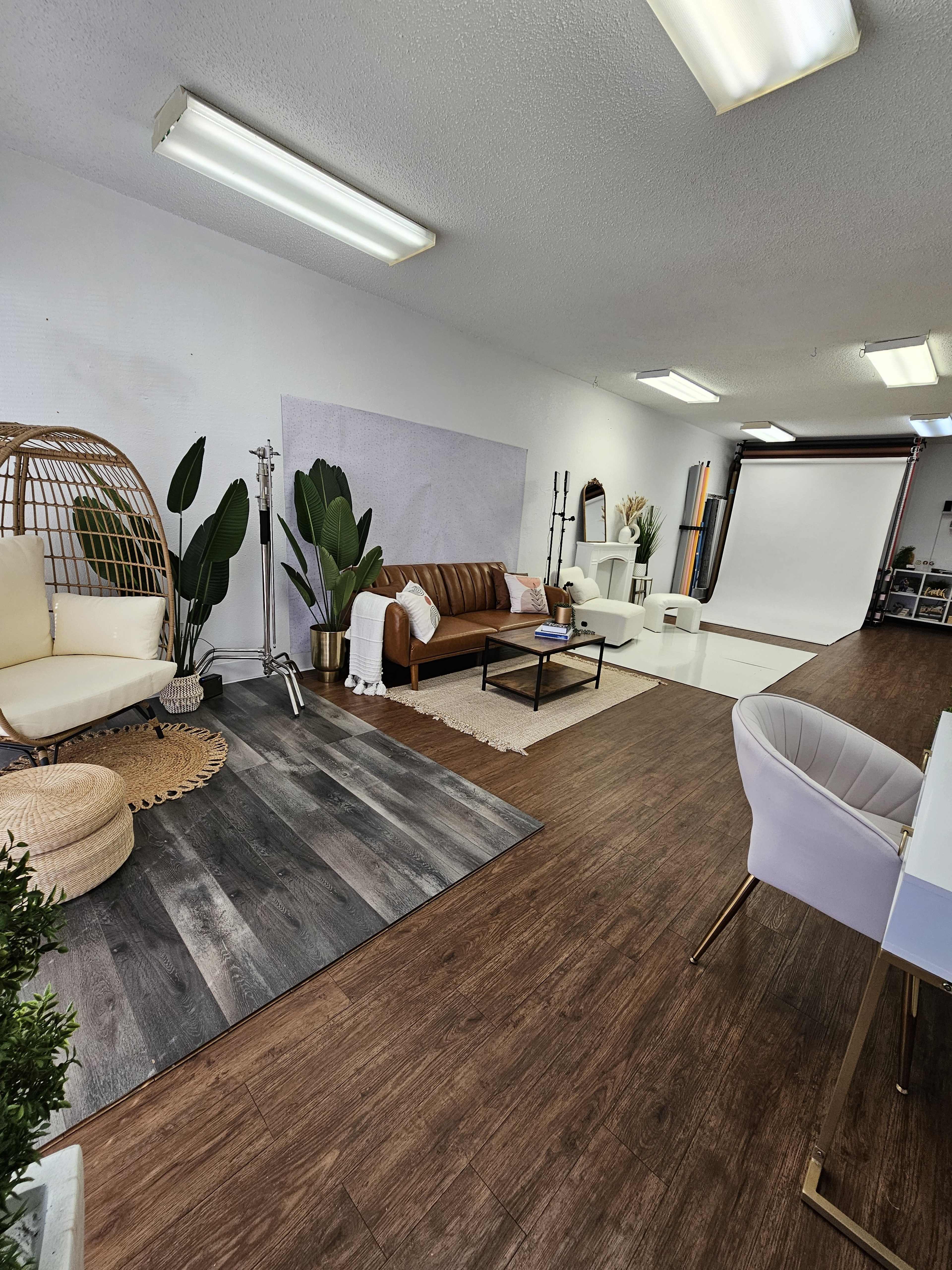 A spacious photography studio featuring a brown leather sofa, a light-colored hanging chair, large plants, and a backdrop area with studio lighting equipment.