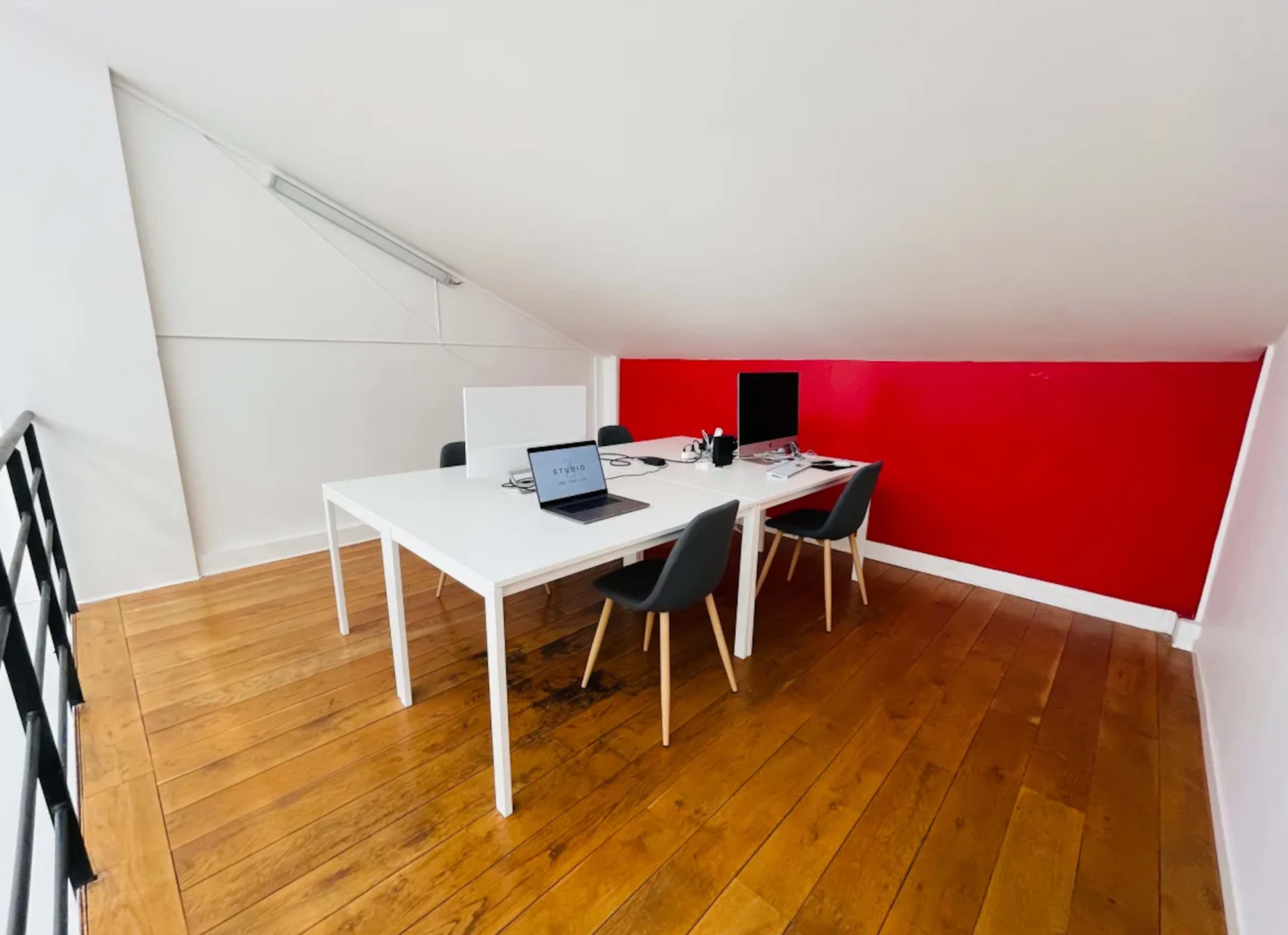 A minimalist office space with two desks, one laptop, and a monitor, set against a red accent wall.