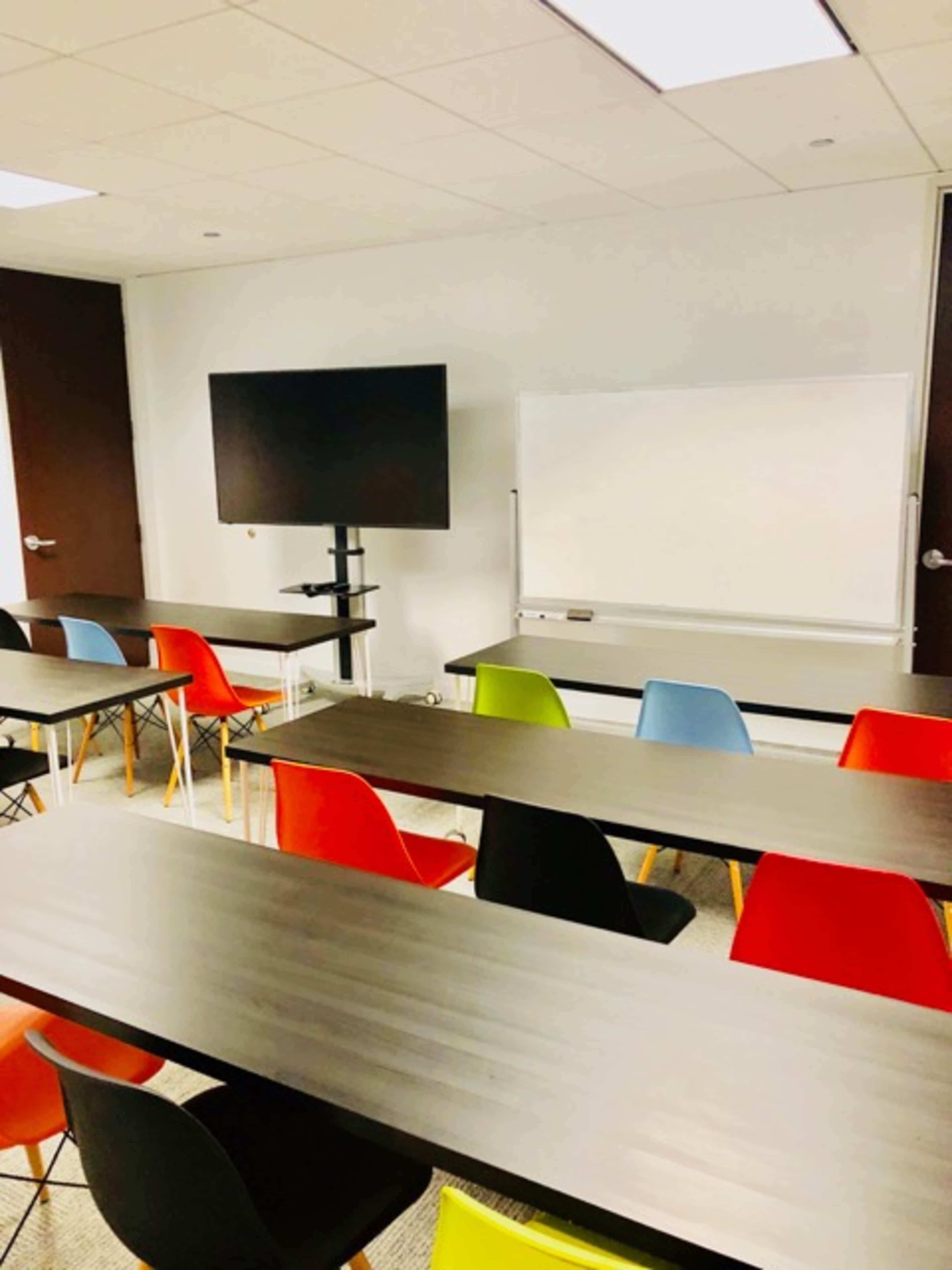 An empty classroom with several tables and colorful chairs arranged in front of a whiteboard and a television monitor.