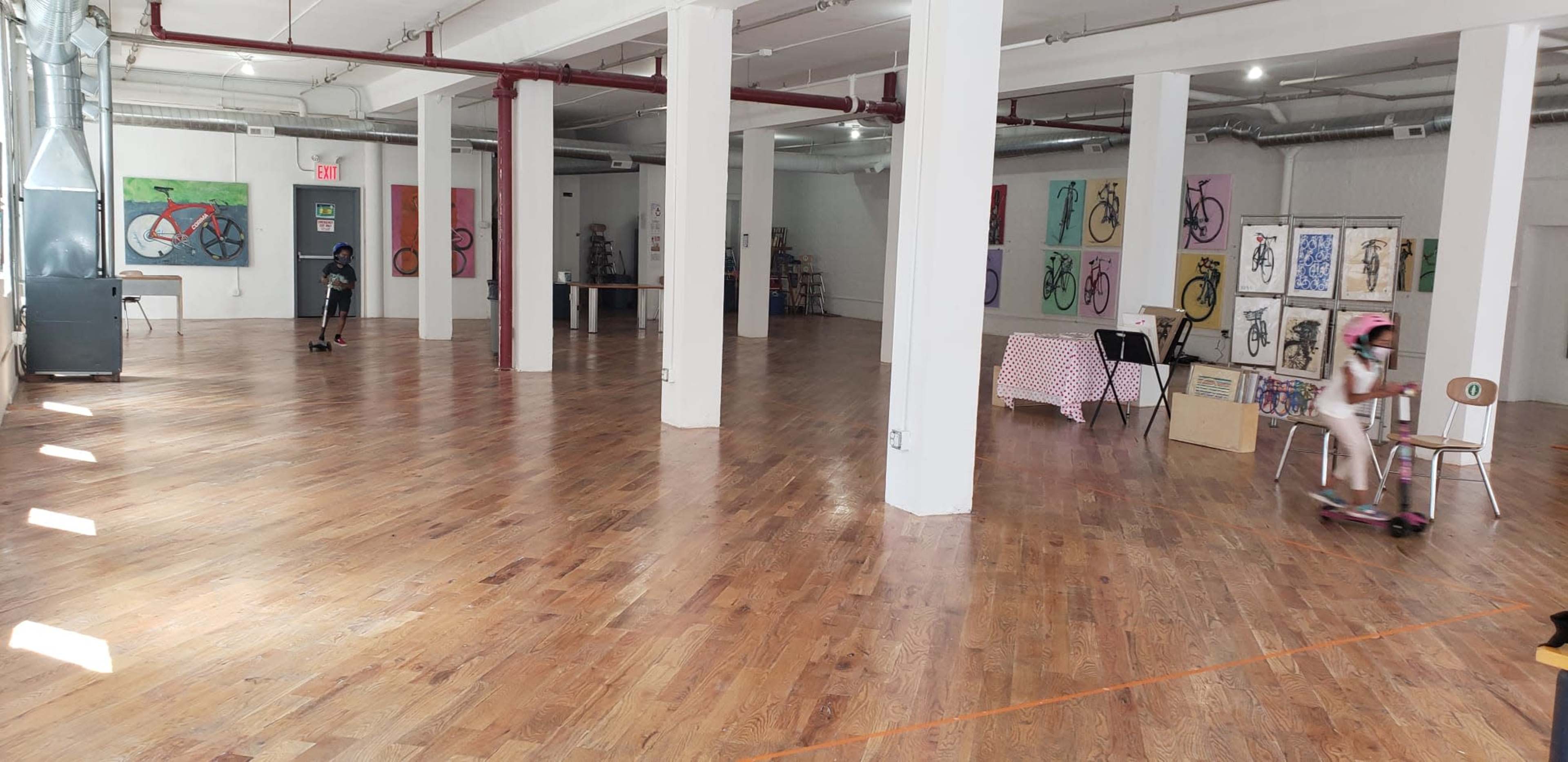 The image shows an open studio space with wooden floors, featuring several artworks on the walls and a child on a scooter.
