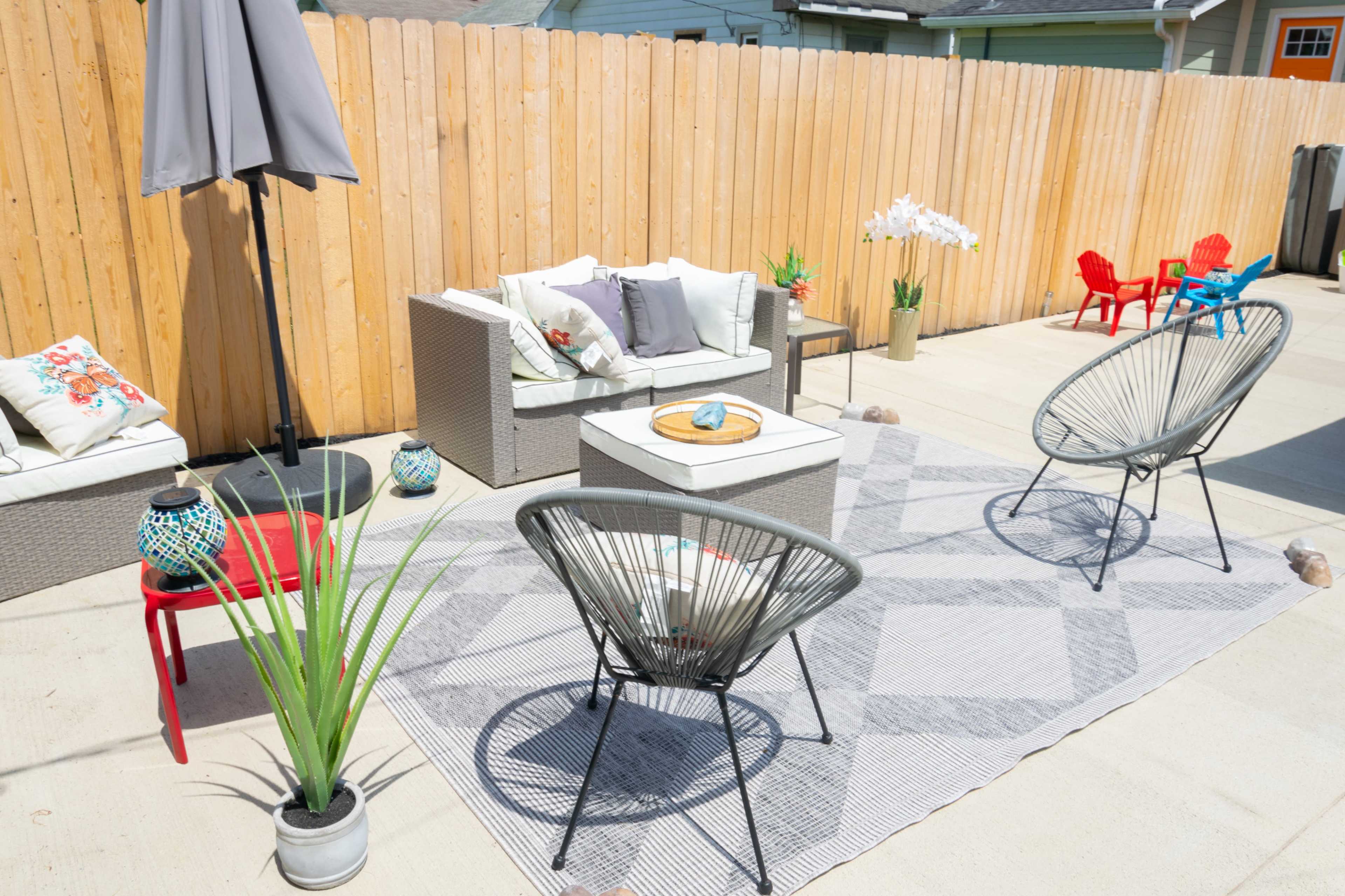 A patio area features a sectional sofa, two circular chairs, decorative plants, and a central rug, surrounded by a wooden fence.
