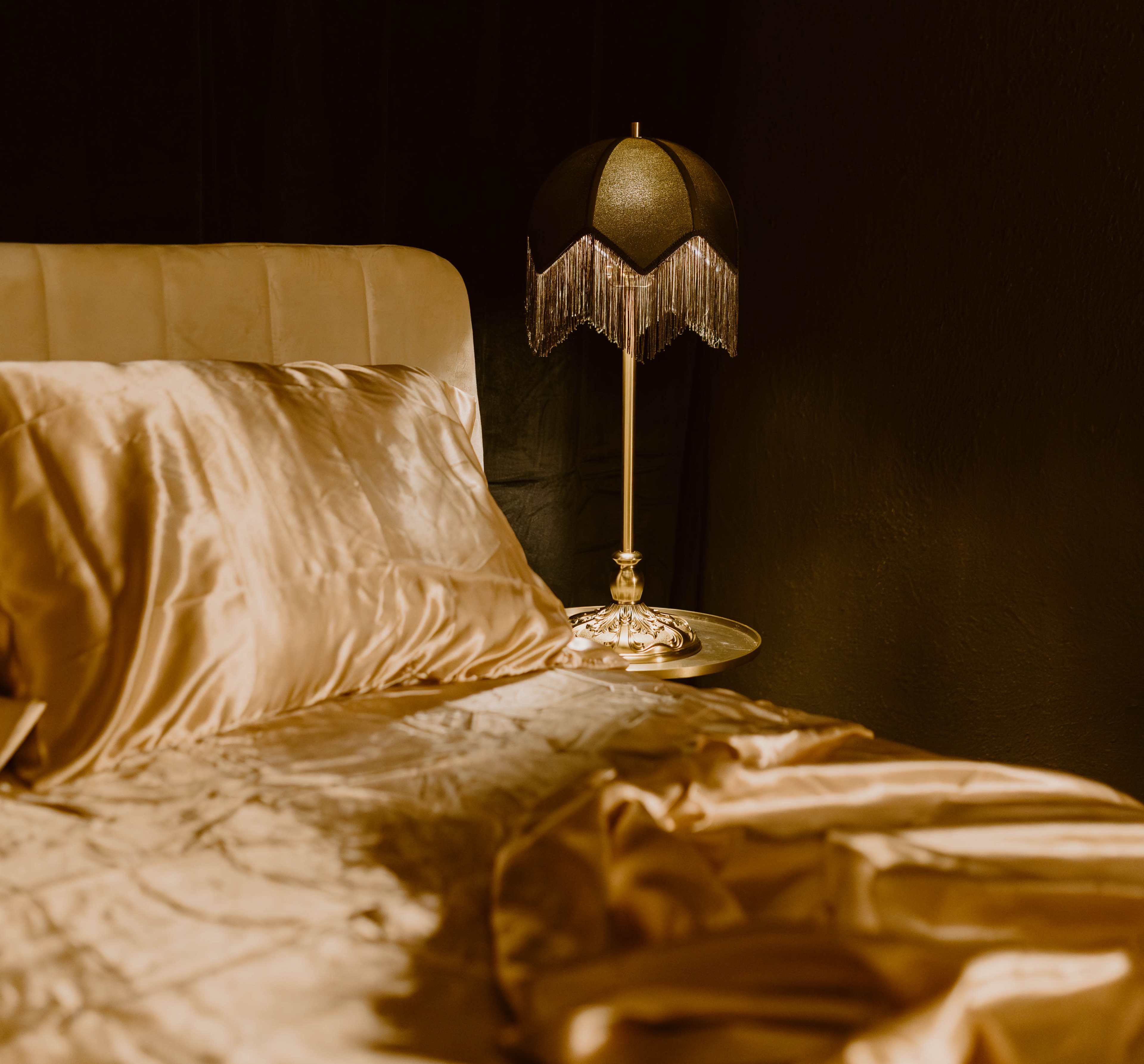 The image shows a neatly arranged bed with golden satin sheets next to a decorative lamp with a fringed shade.