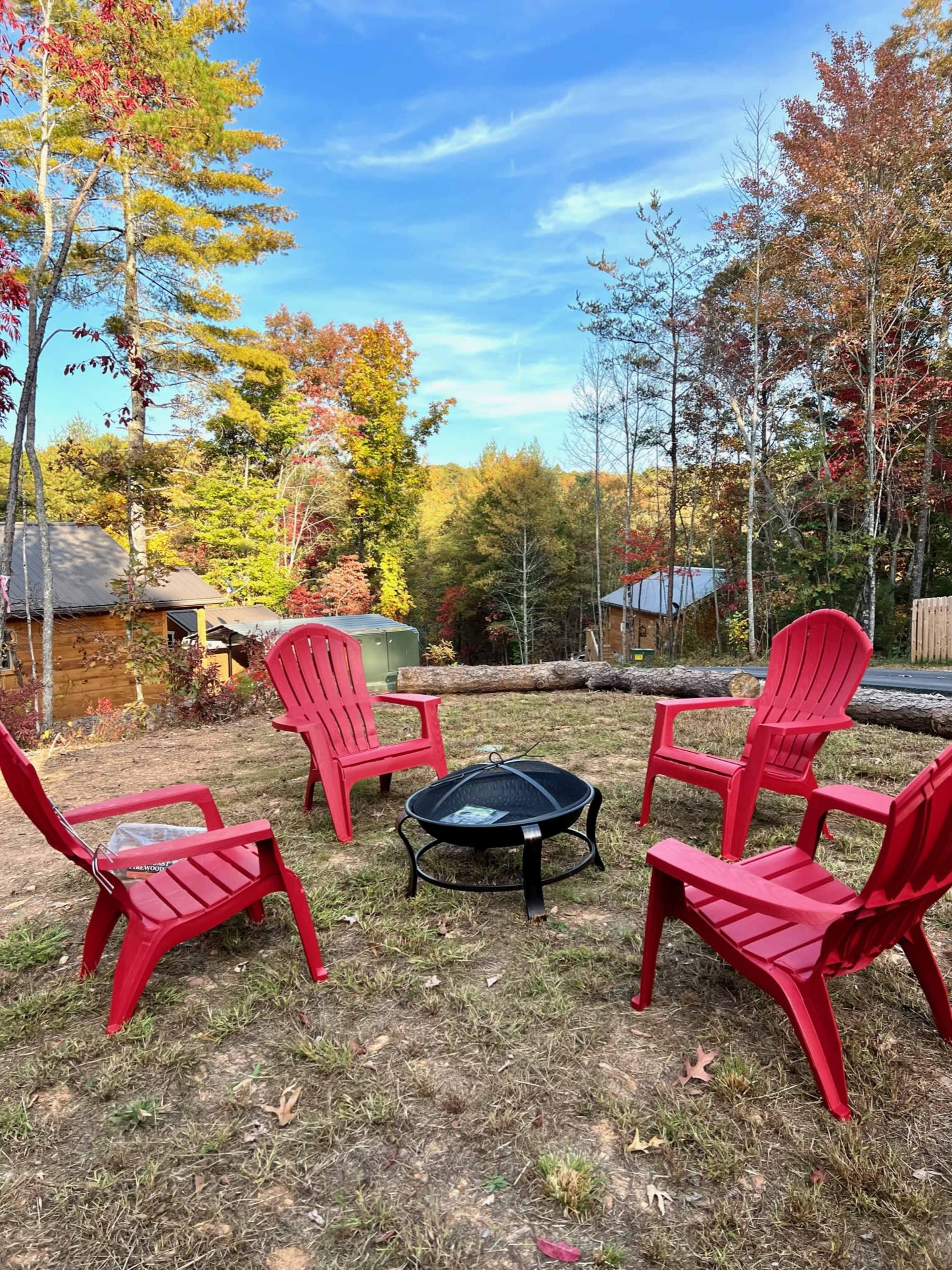 A circular arrangement of red chairs surrounds a black fire pit on a grassy area, with trees displaying autumn colors in the background.
