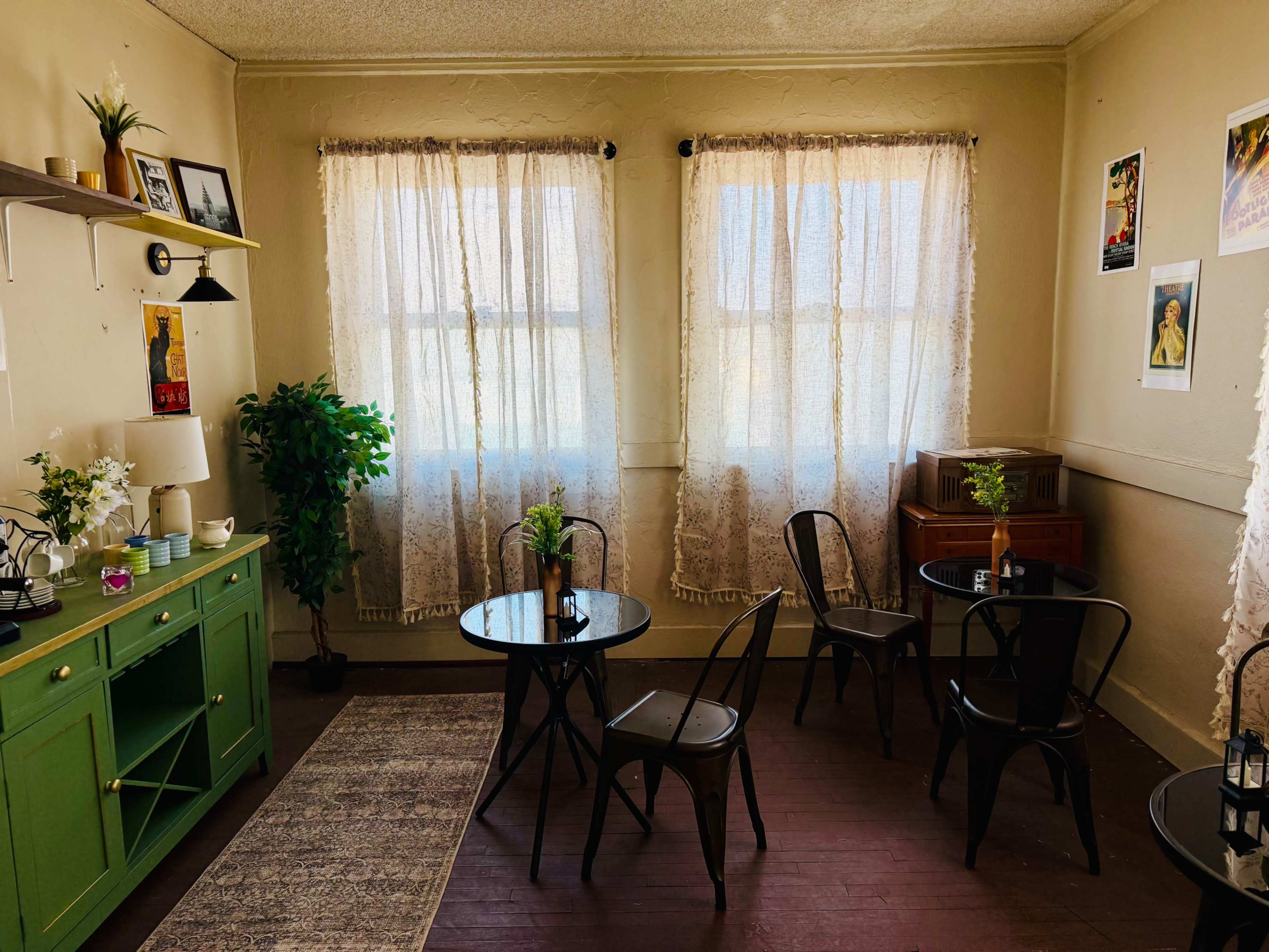 The image shows a cozy room with two large windows covered by sheer curtains, a green console table, two small round tables with black chairs, and a decorative plant in the corner.