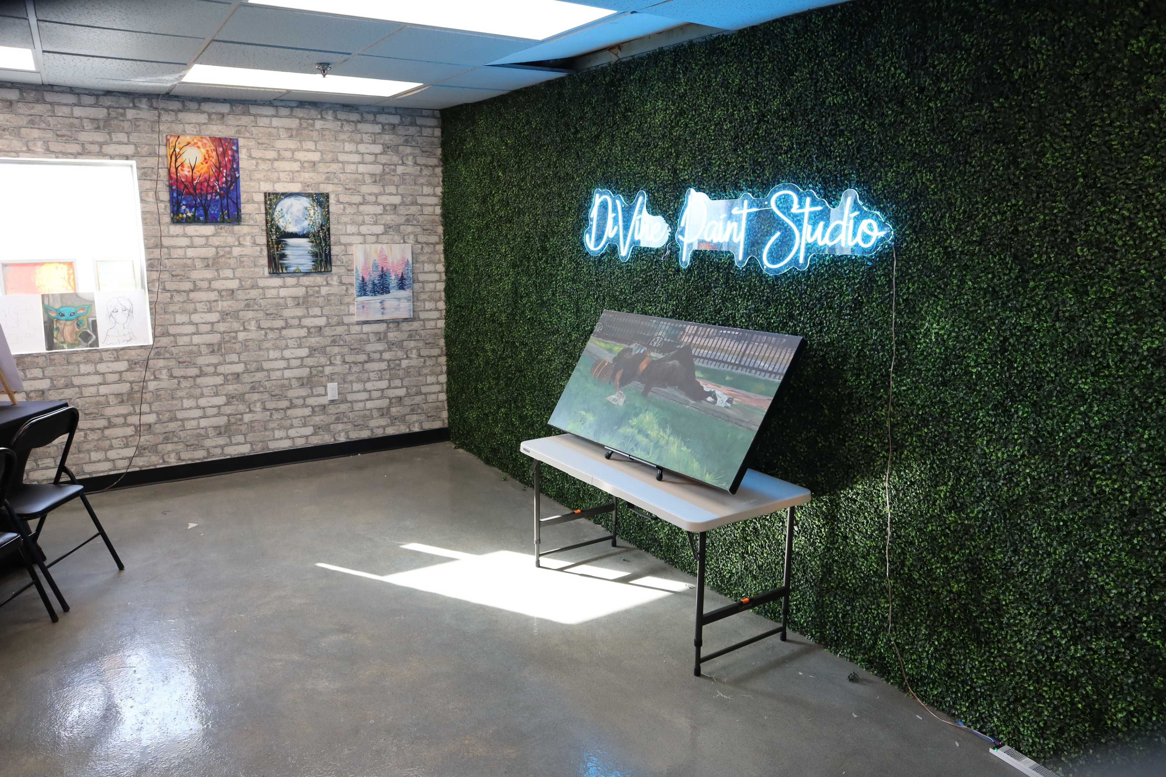 The scene shows a cozy art studio featuring a green, textured wall, a TV on a table, and several artworks displayed in frames.