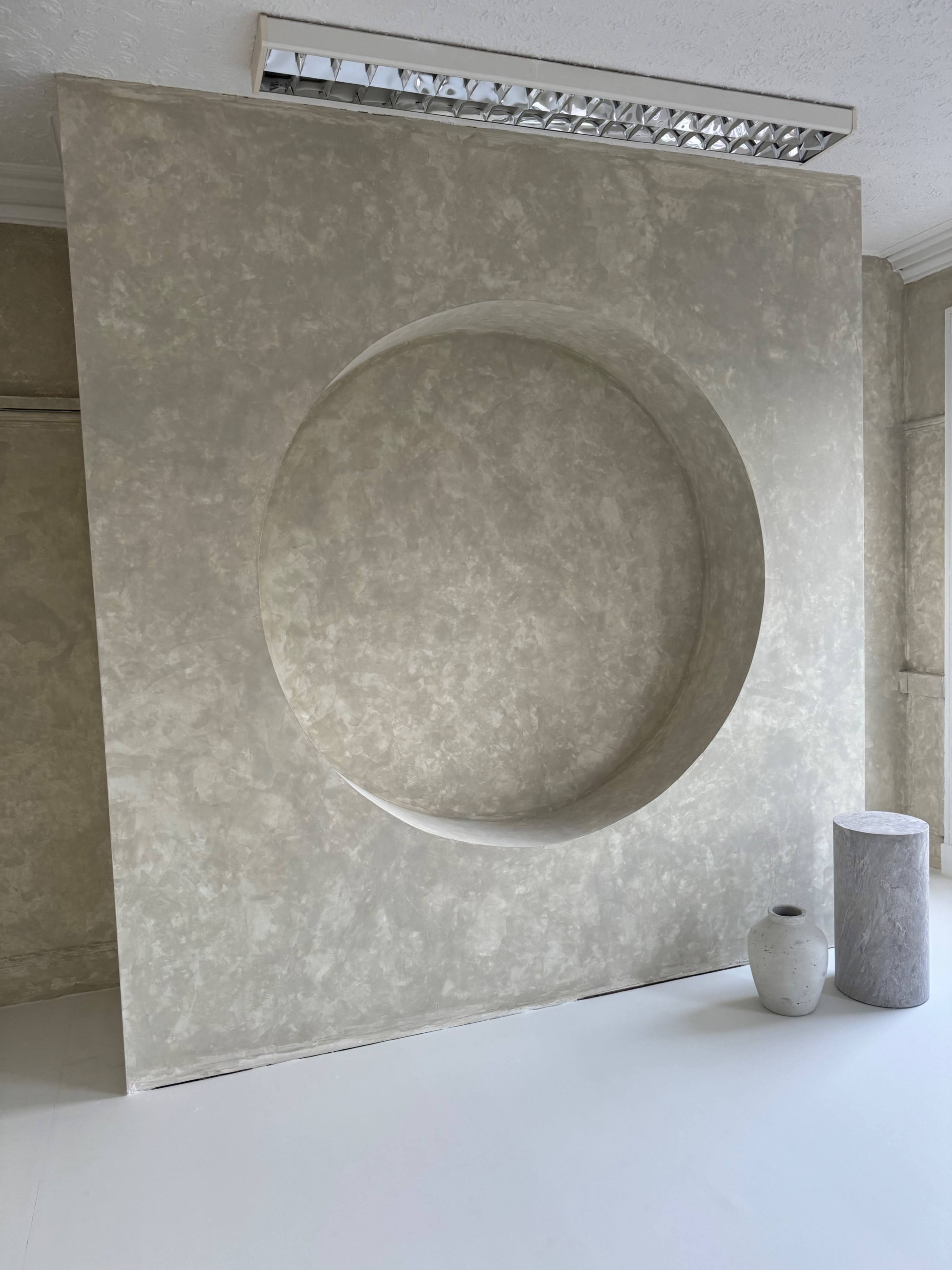 The image shows a textured wall with a large circular cutout, accompanied by a vase and a cylindrical stone object on a light-colored floor.