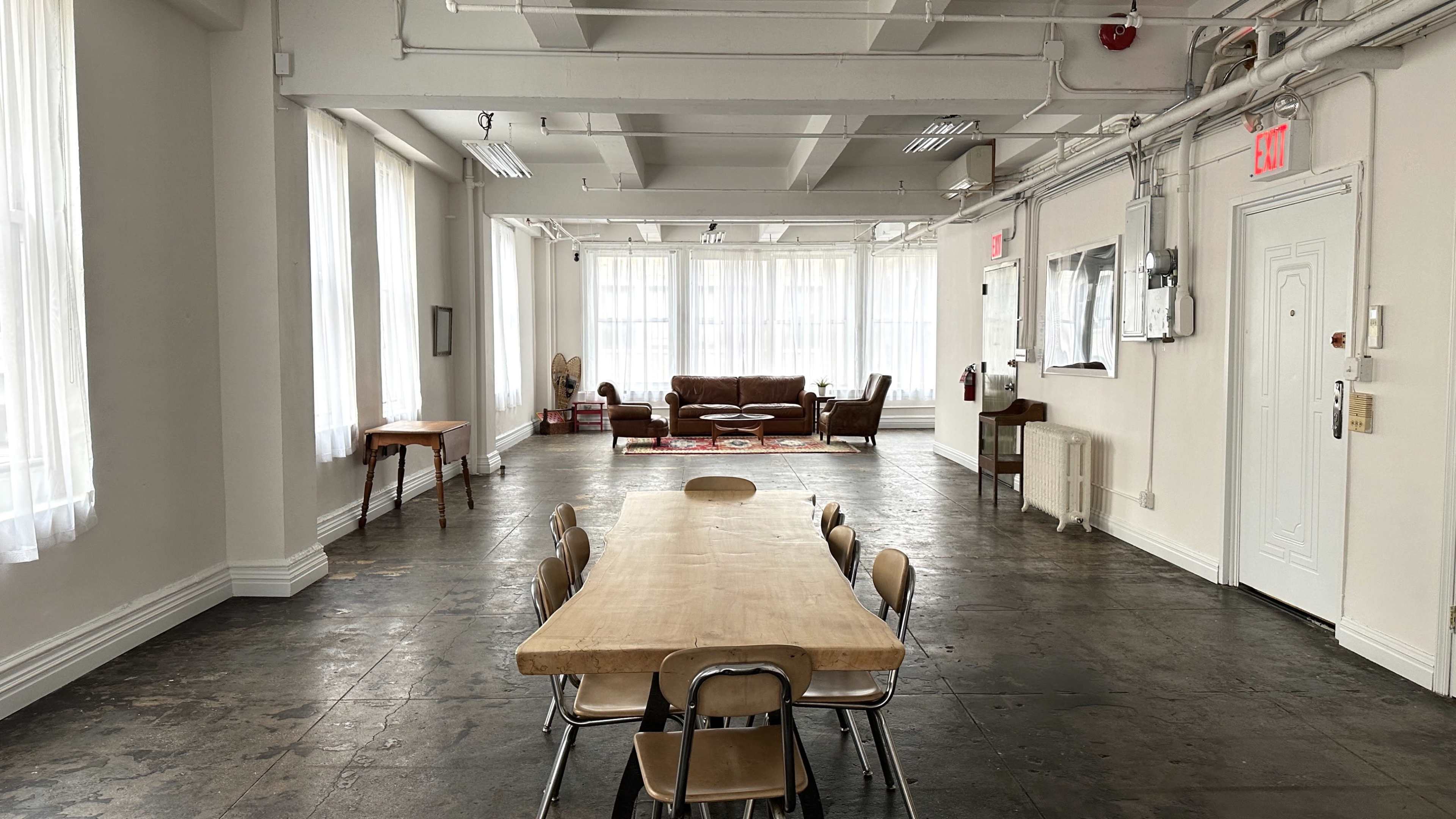 A spacious, well-lit room features a long wooden table surrounded by chairs, with a seating area and large windows in the background.