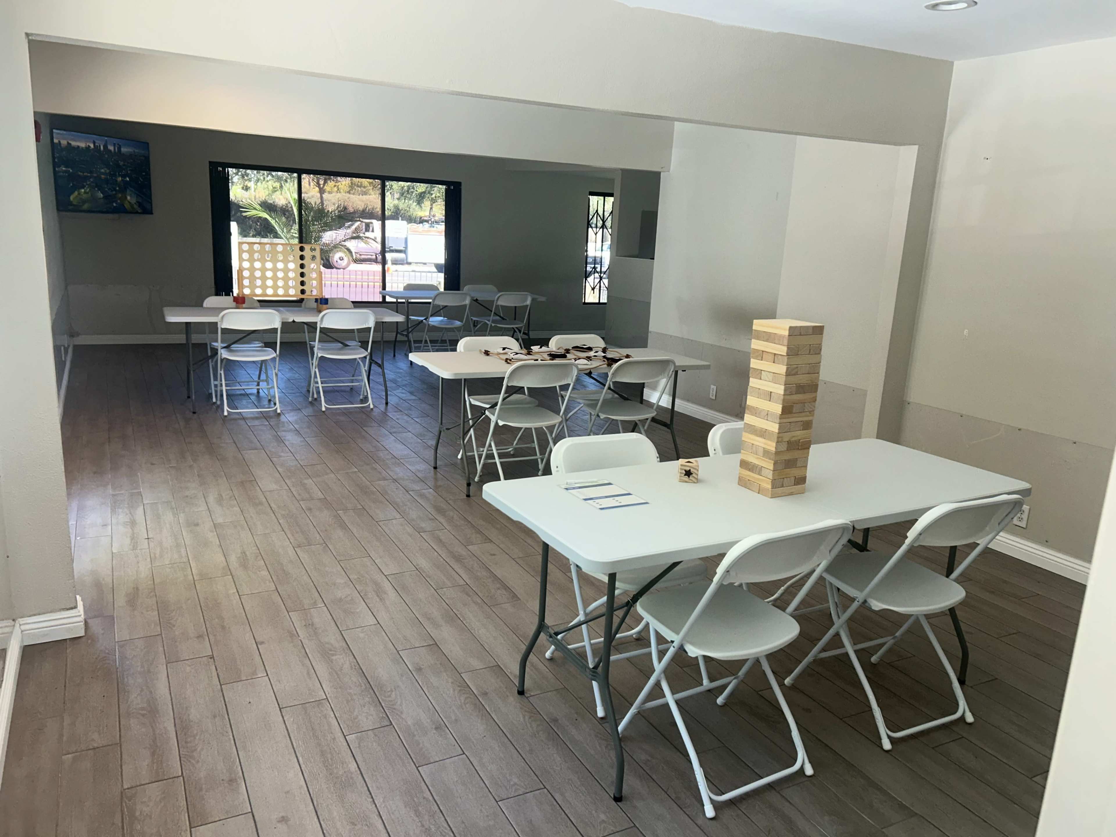 The image shows a spacious room with several white folding tables and chairs arranged for gatherings, with a large window allowing natural light and a game of Jenga prominently displayed in the center.