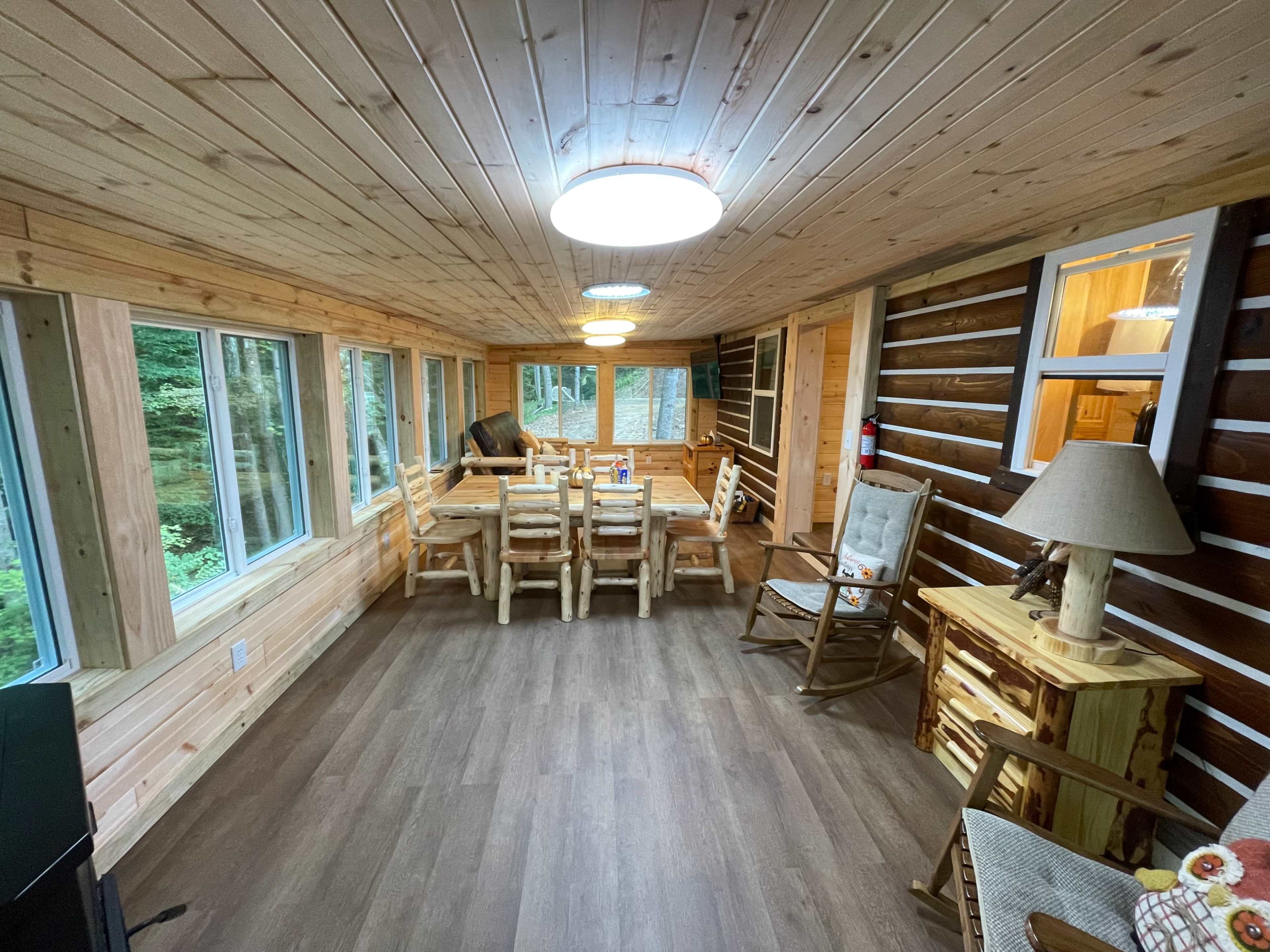 A rustic dining area features wooden walls and ceilings, large windows, a wooden table with six chairs, and various wooden furnishings.