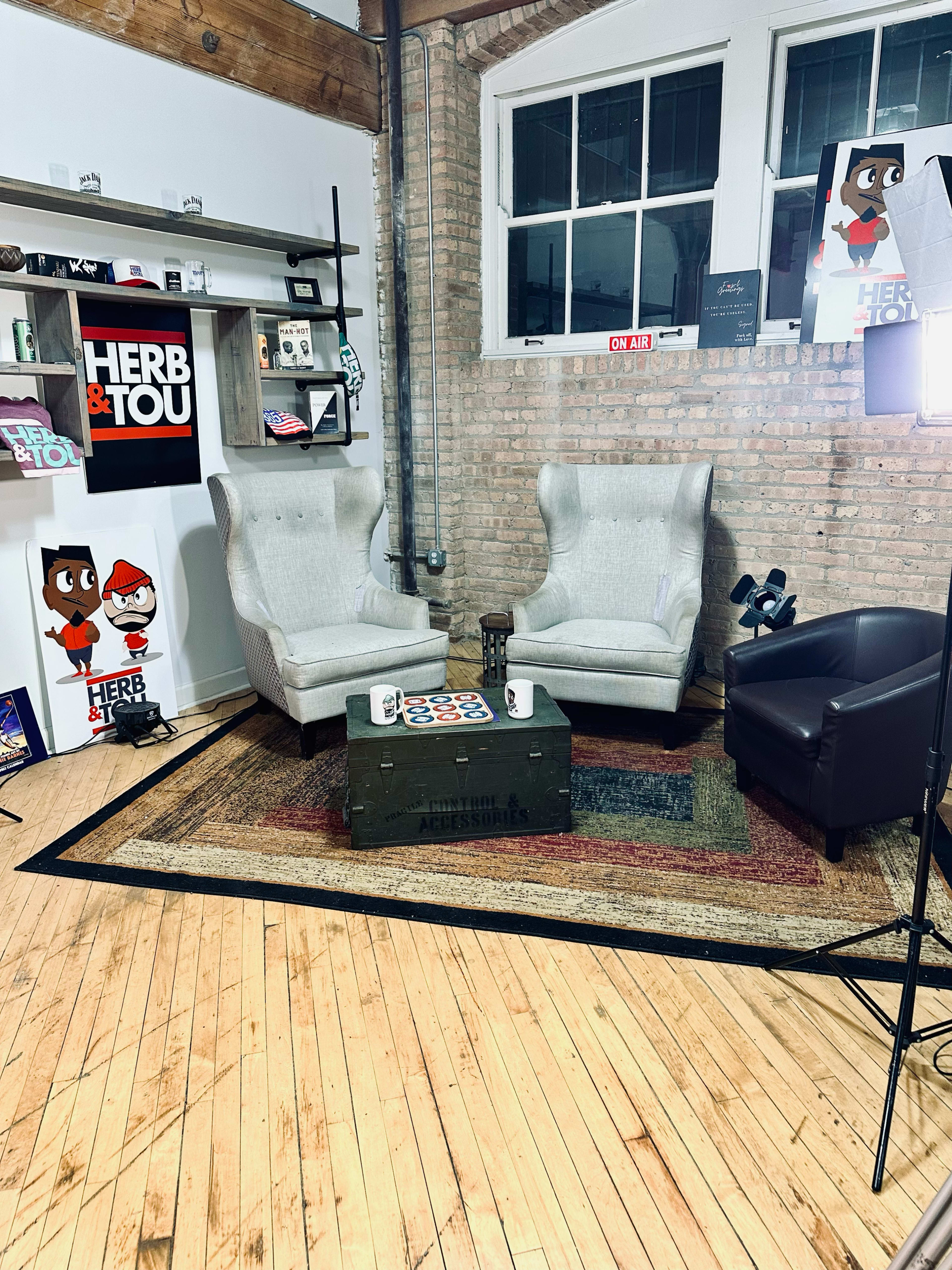 The image depicts a cozy studio setting featuring two wingback chairs, a dark leather chair, a coffee table, and various decorations on the walls, including artwork and a shelf filled with items.