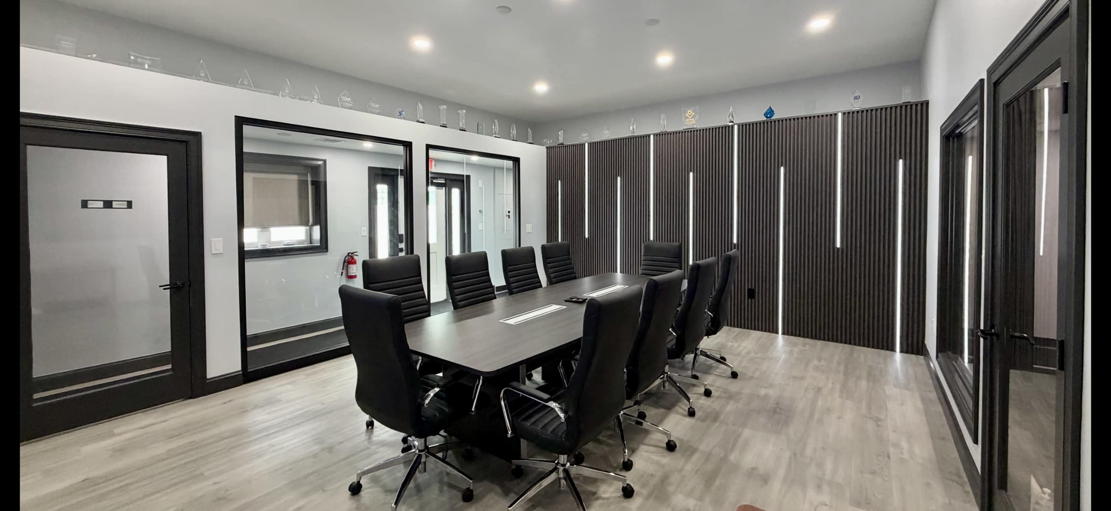 The image shows a modern conference room with a large table surrounded by black chairs, accented by wall-mounted trophies and a glass partition.