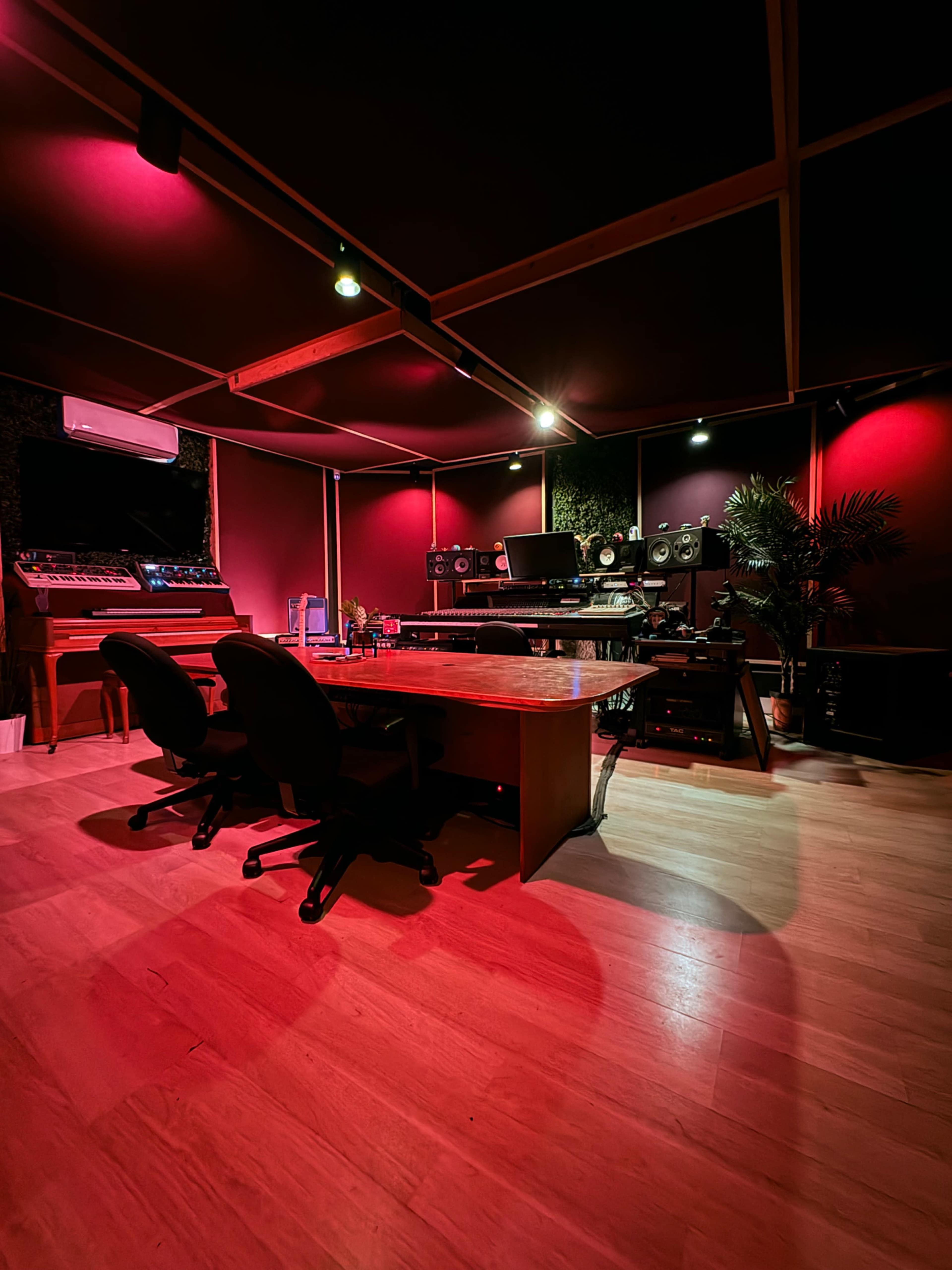 The image shows a music studio with red lighting, featuring a large wooden table, two black chairs, and various audio equipment against the walls.