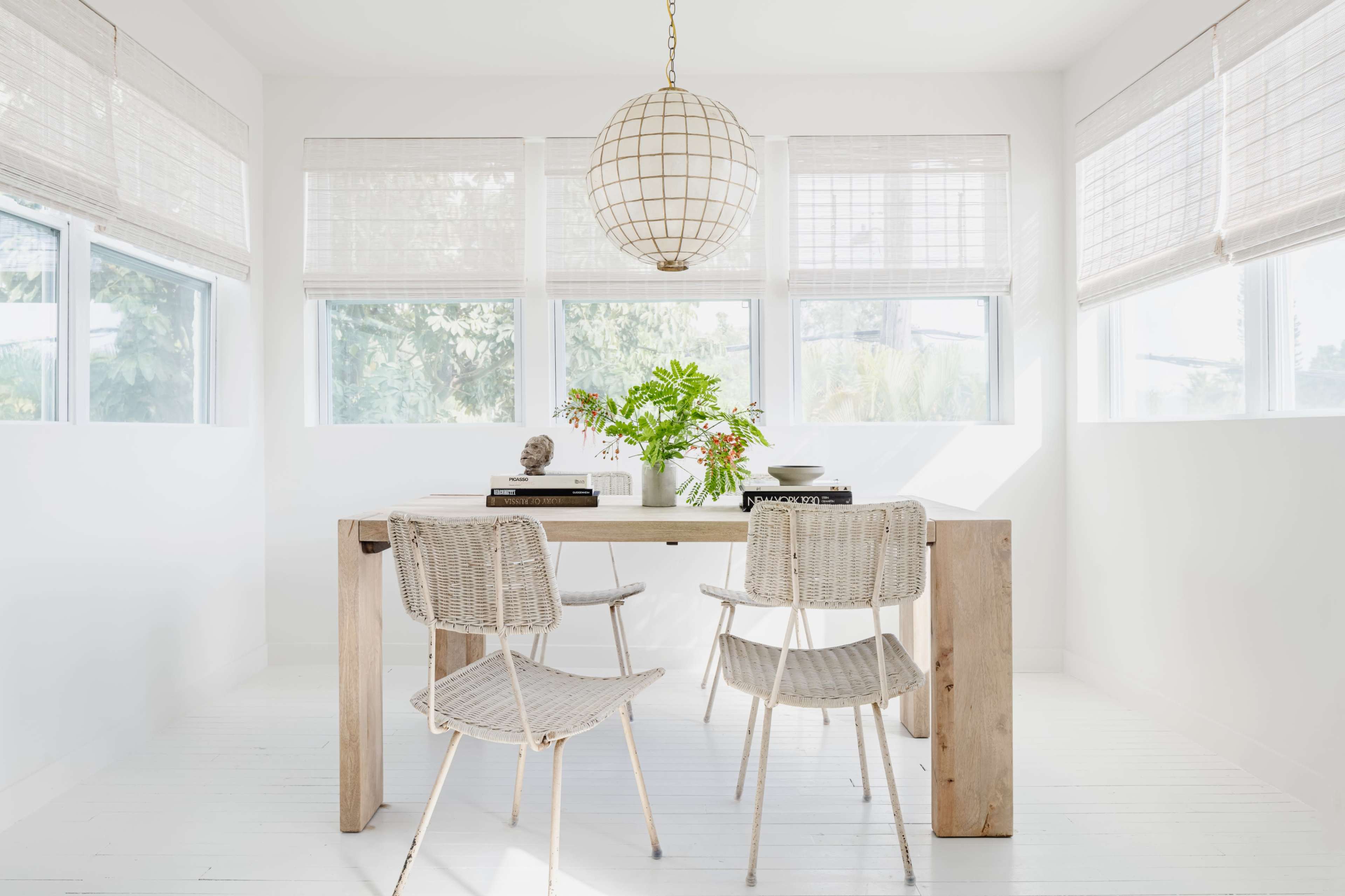 A minimalistic dining area features a wooden table surrounded by two woven chairs and a pendant light, with large windows allowing natural light to fill the space.