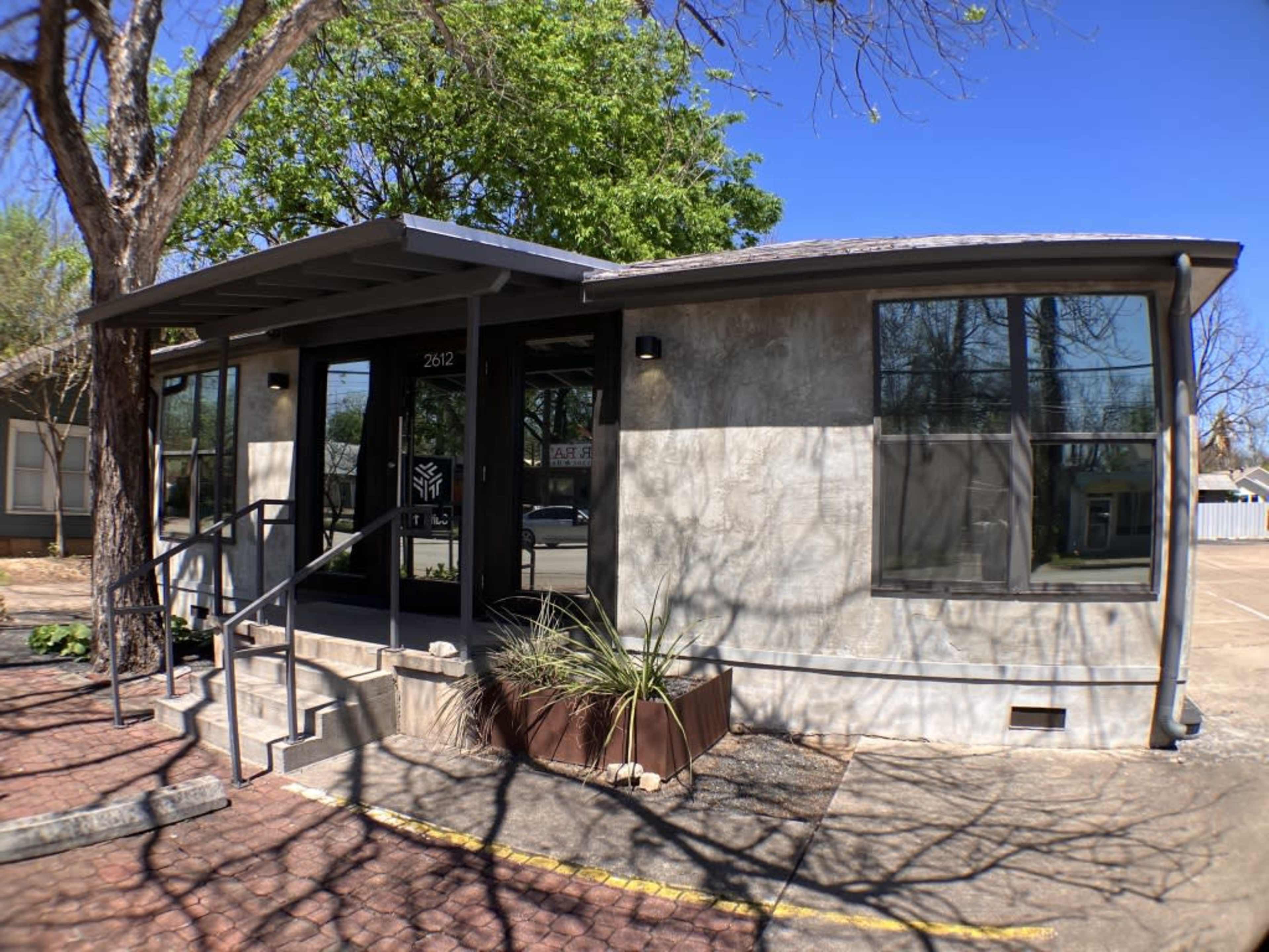A modern building with large windows, a flat roof, and a landing with steps in front, surrounded by trees and landscaping.