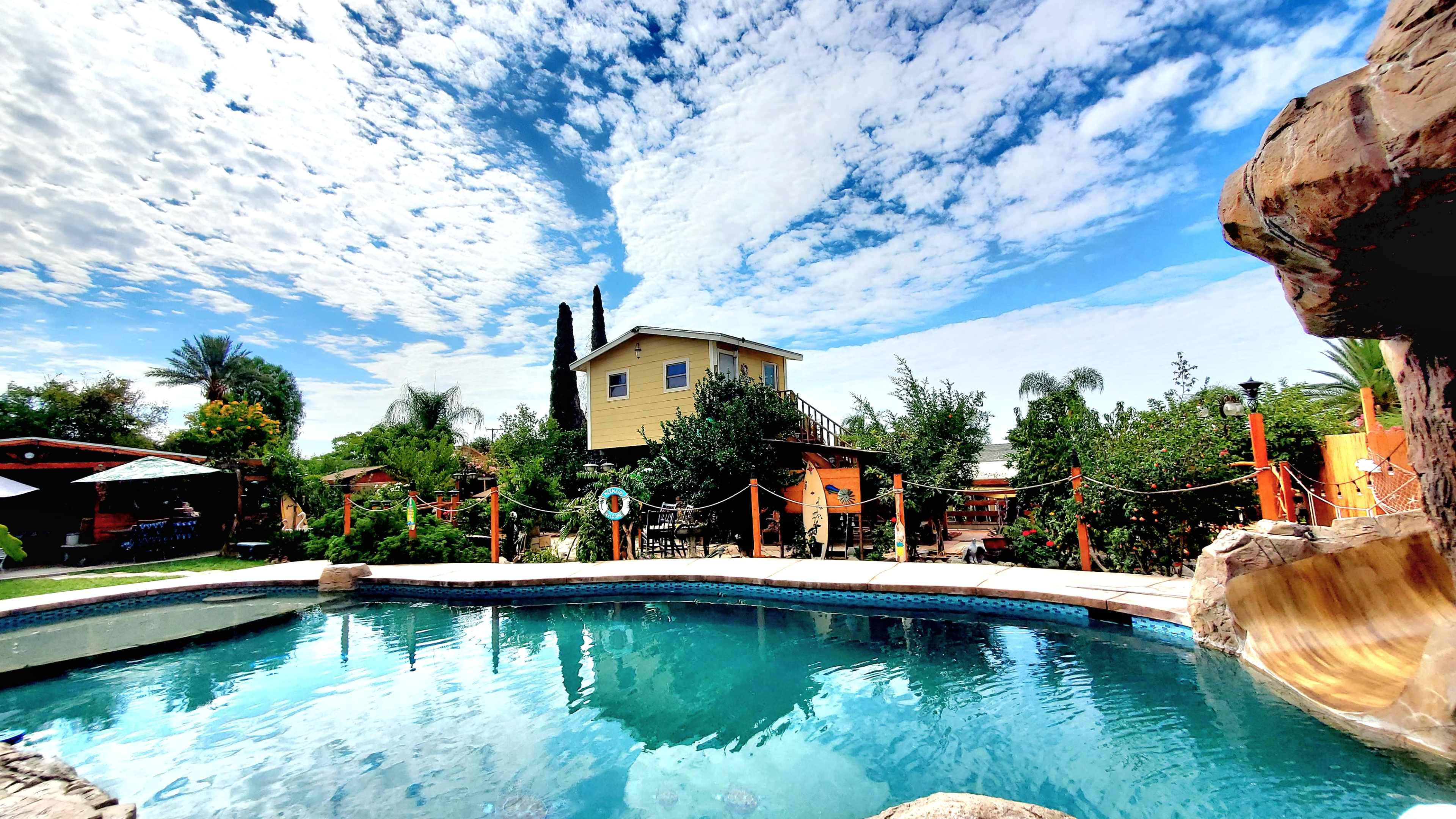 The image shows a clear swimming pool surrounded by lush greenery and a two-story house in the background under a partly cloudy sky.