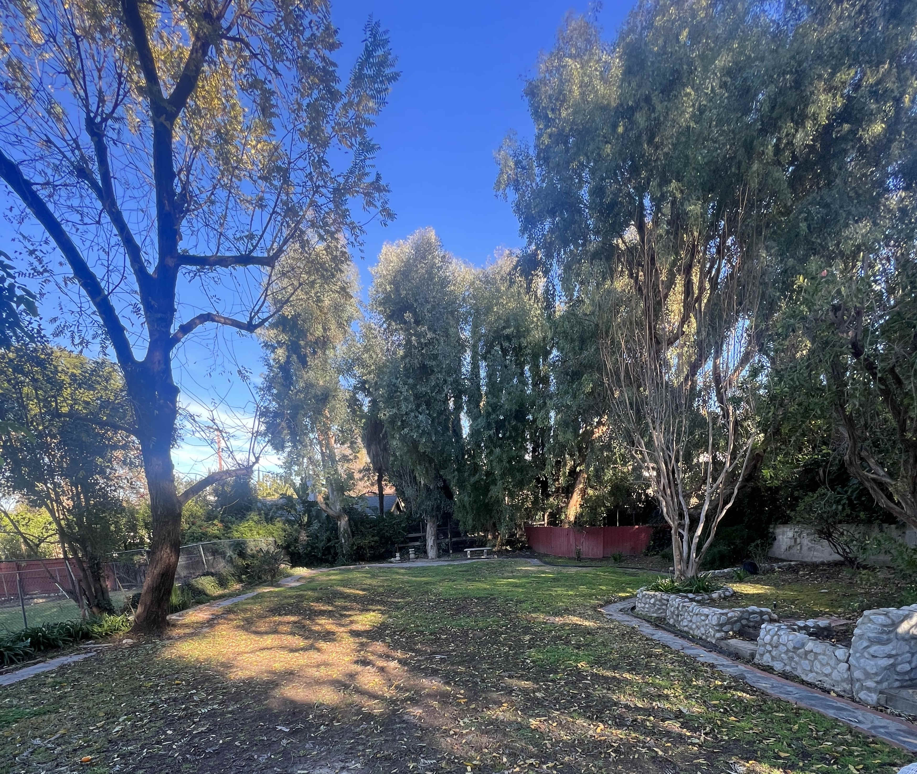 A grassy area is surrounded by tall trees, with a stone path and a red structure in the background.