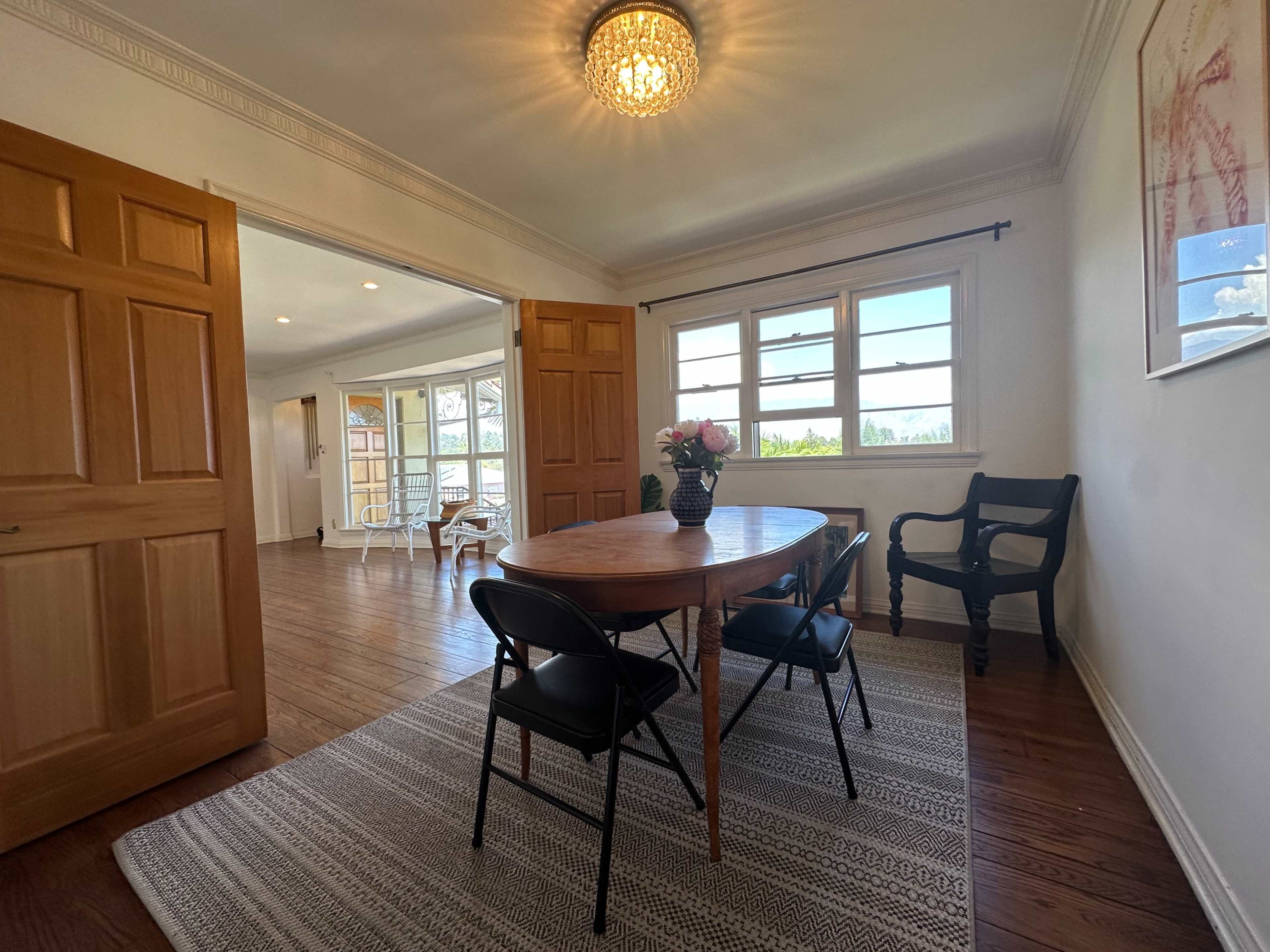A dining area features a wooden table with three black chairs and a vase of flowers, adjacent to an open space with large windows.