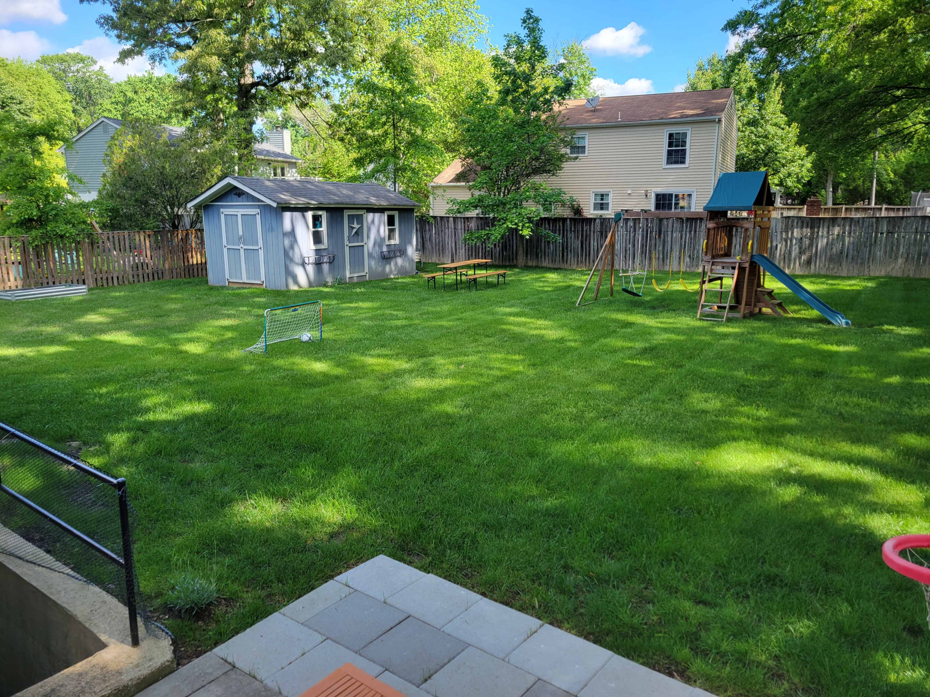 The image shows a grassy backyard featuring a wooden playset, a small shed, a picnic table, and a soccer goal.
